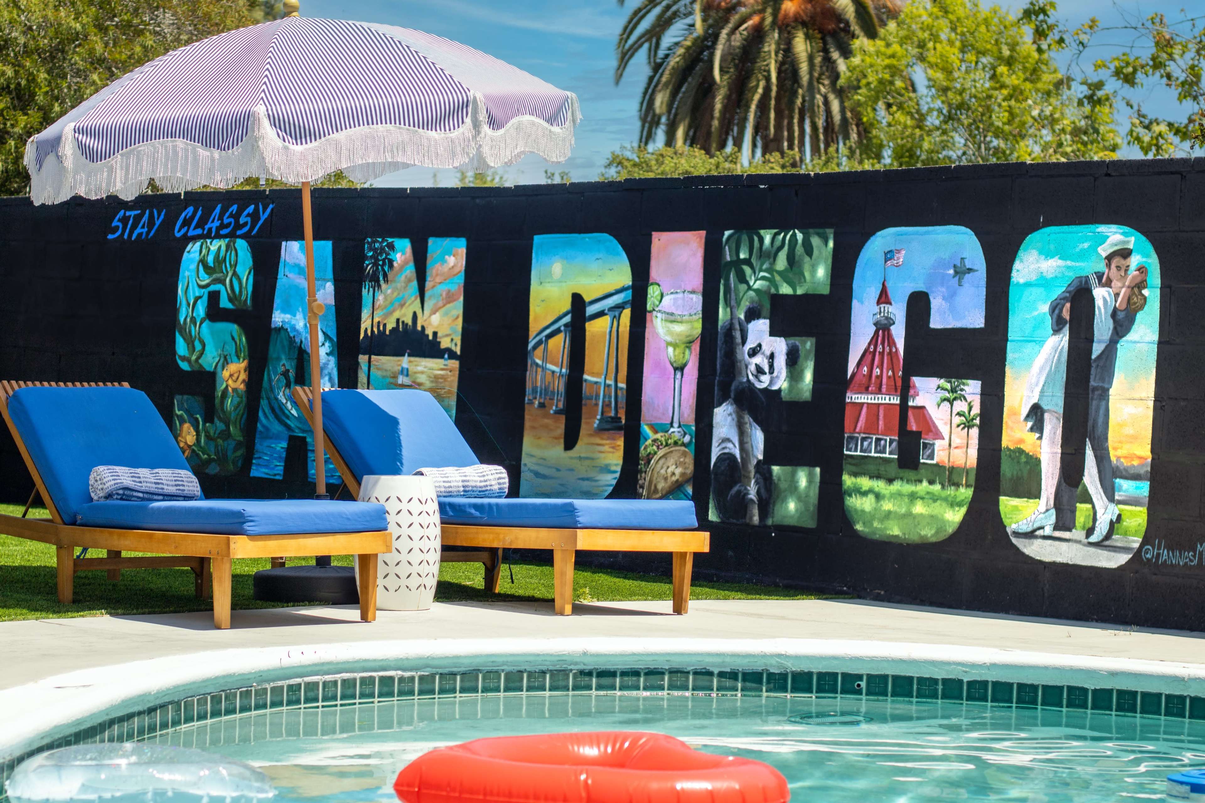 A swimming pool with a red floatation device is surrounded by two lounge chairs and an umbrella, against a wall featuring a colorful mural depicting the words "San Diego" and various local scenes.