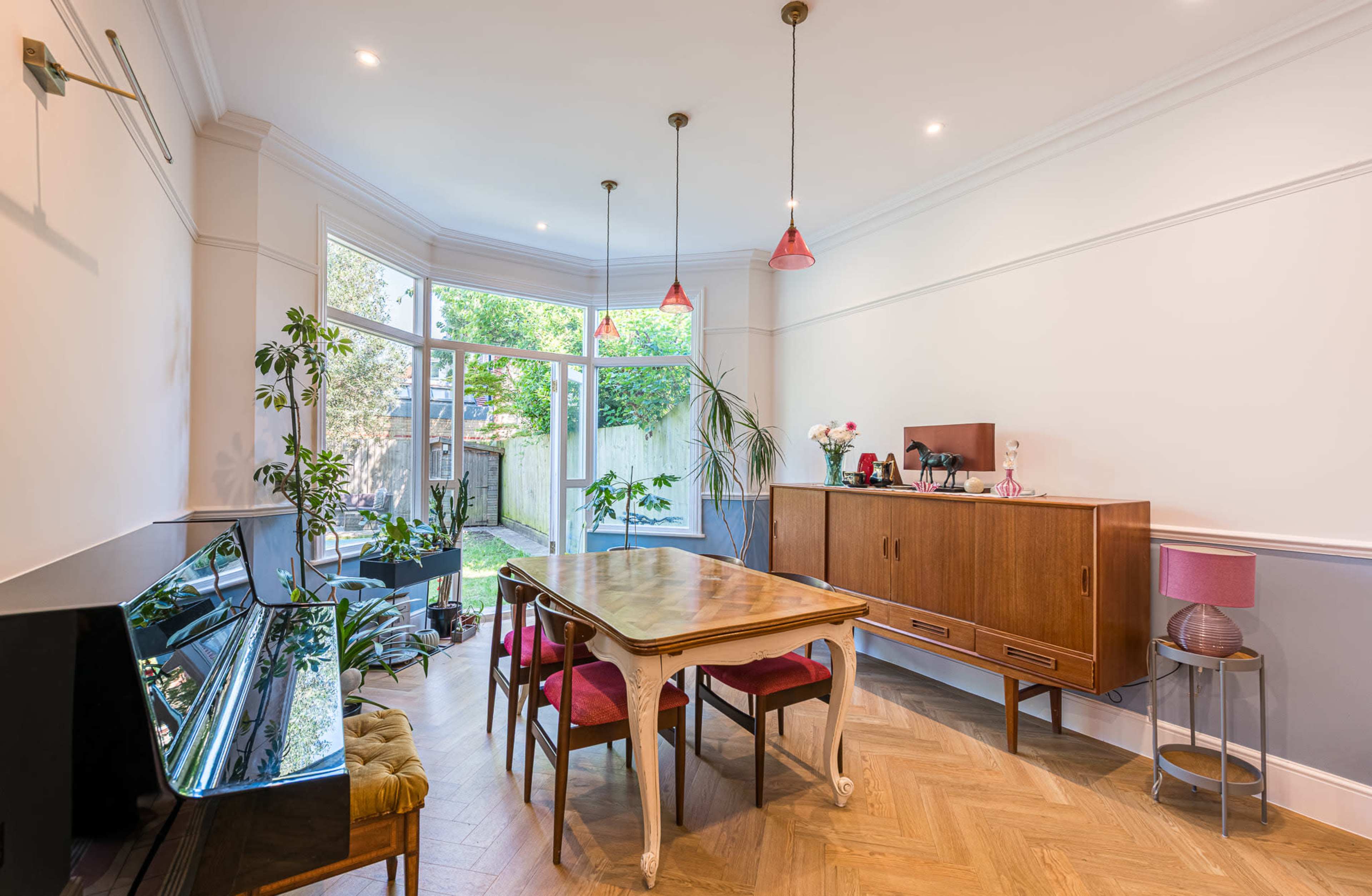 The image depicts a dining area featuring a wooden table surrounded by four chairs, a piano, and large windows that provide access to a garden filled with greenery.