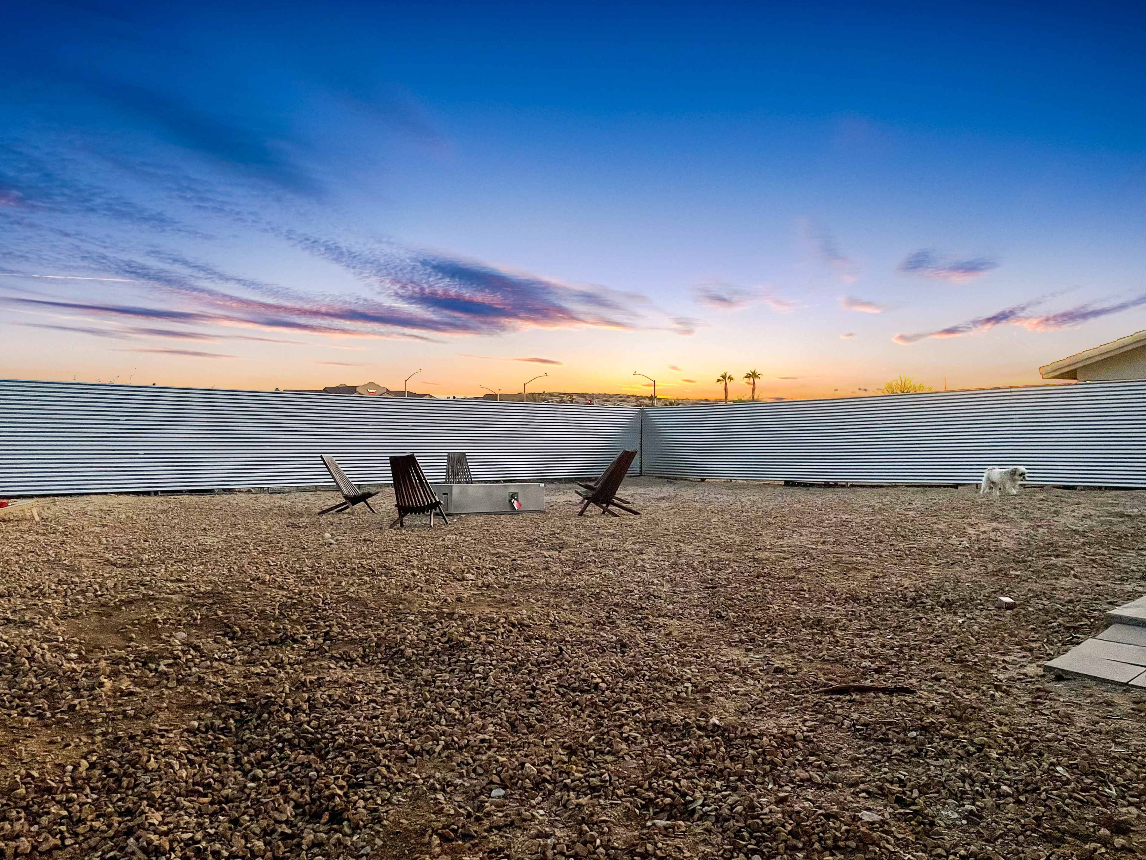 The image shows a gravel-covered yard surrounded by corrugated metal fencing, featuring two wooden chairs and a small fire pit under a colorful sunset sky.