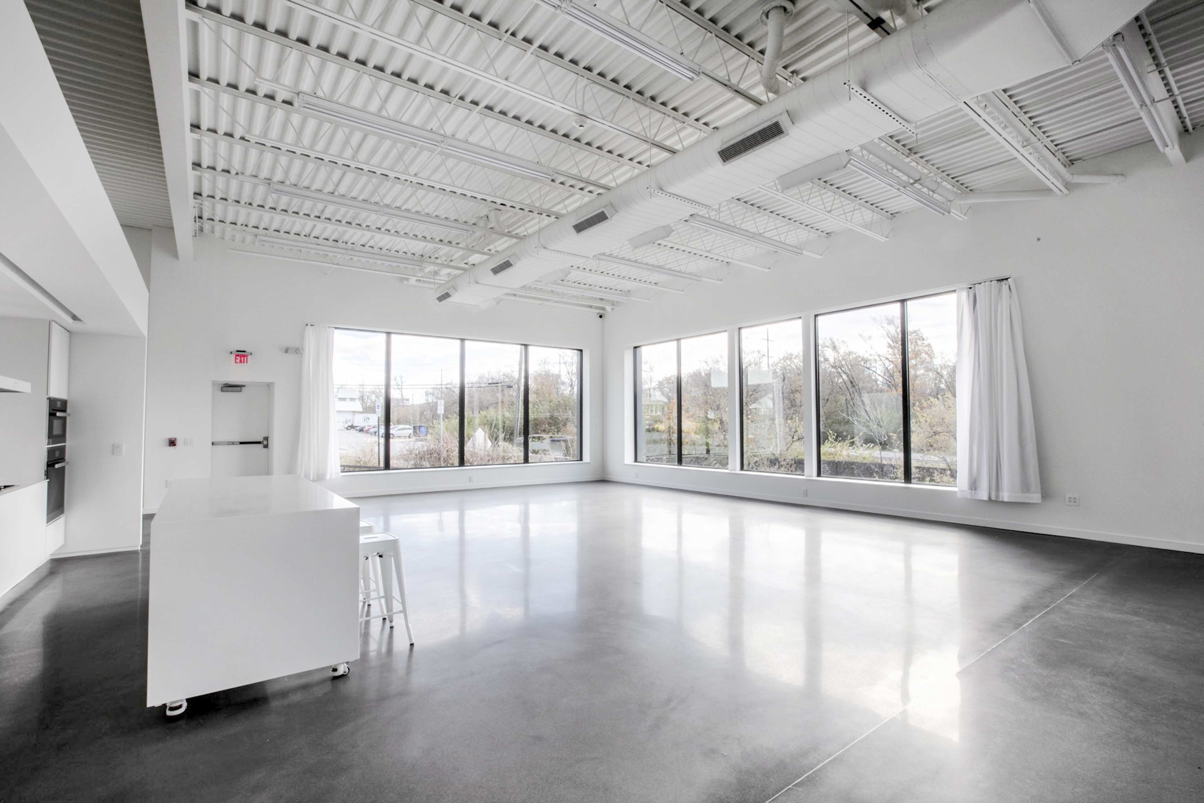 The image shows a spacious, empty room with large windows and a kitchen area featuring a white countertop.