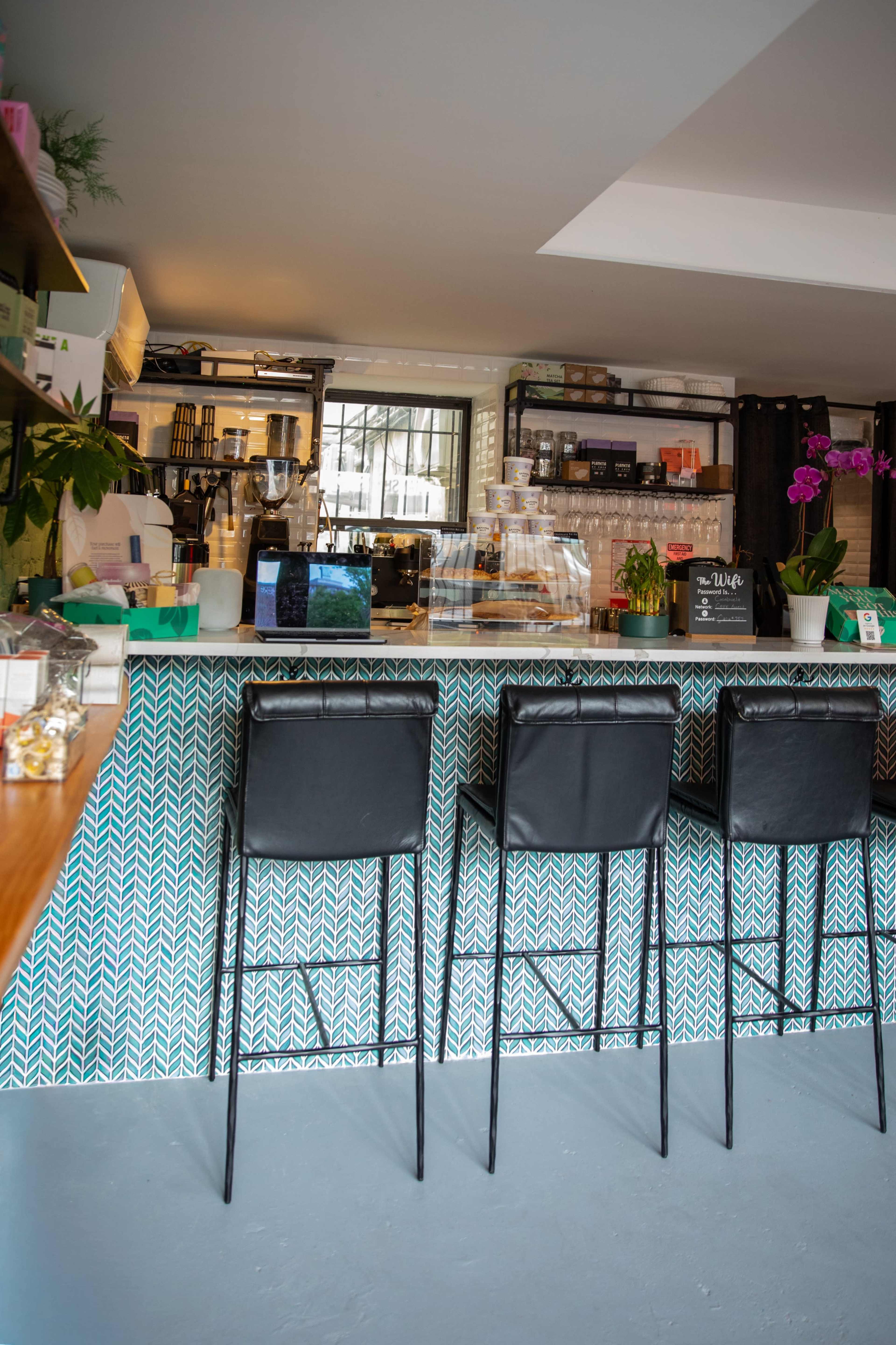 The image shows a modern cafe interior with a patterned bar, three black barstools, and a counter that displays various items and plants.