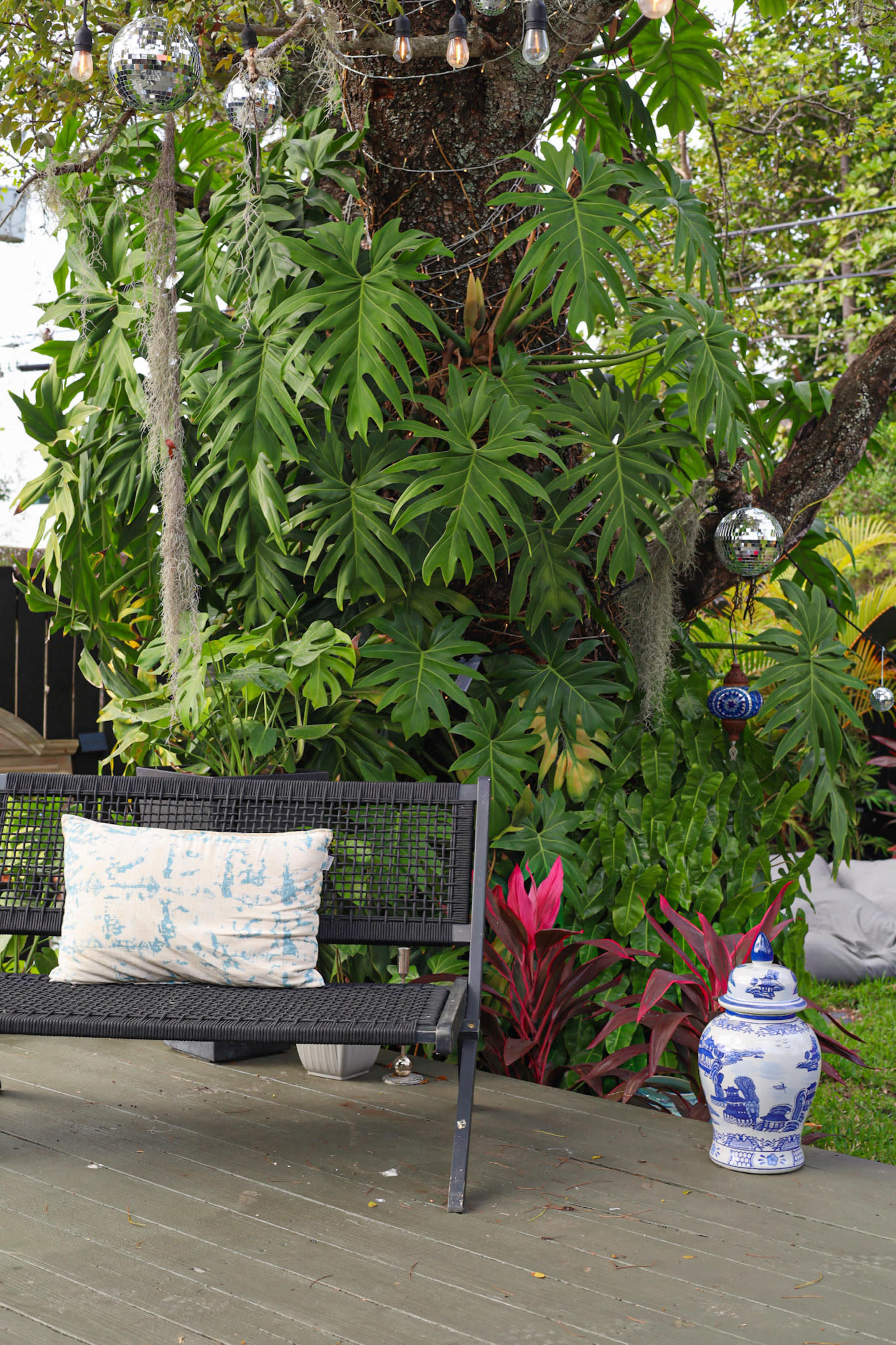 A black bench with a decorative pillow sits on a wooden deck in front of a lush green backdrop featuring large leaves and hanging ornaments.