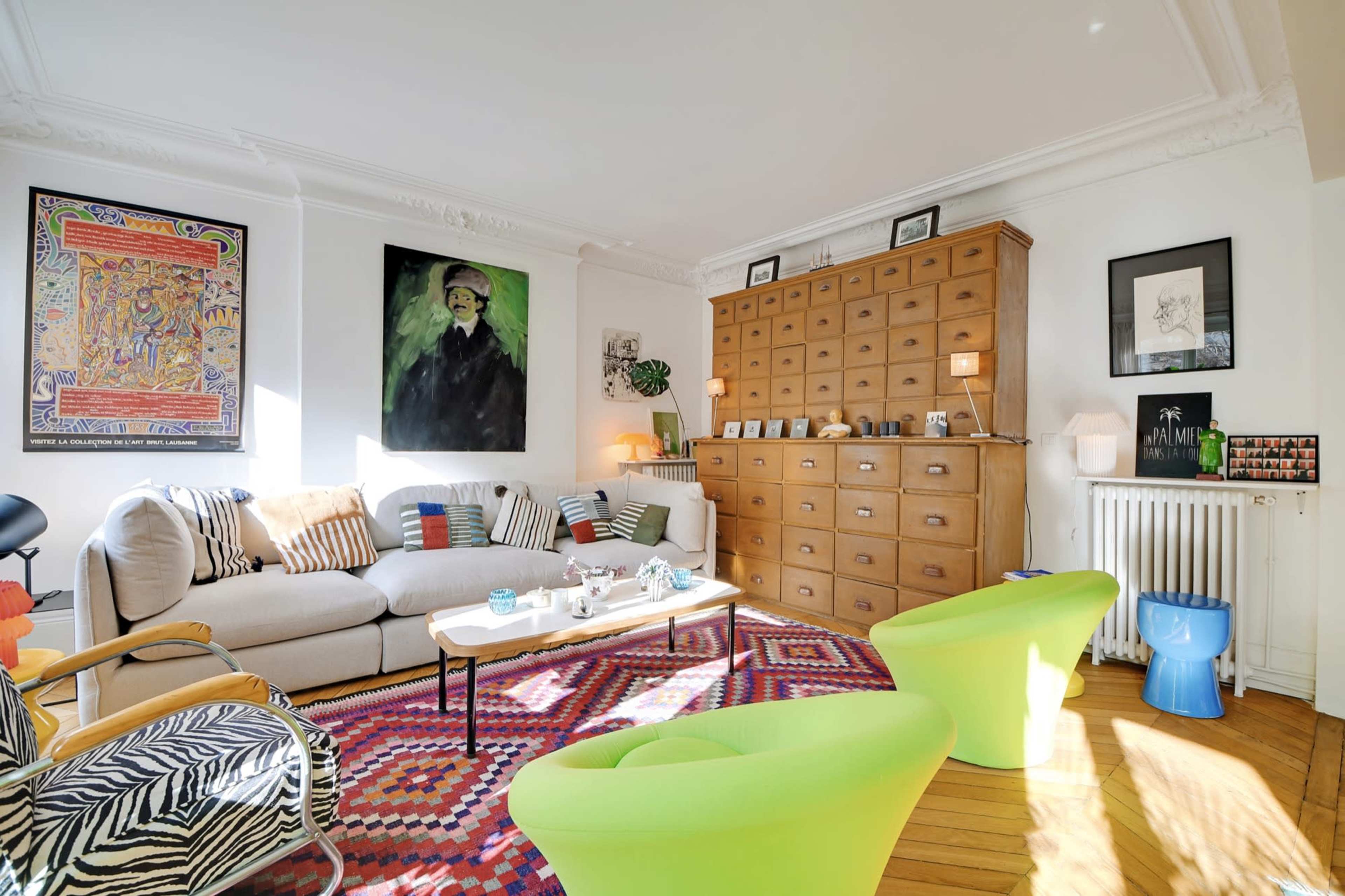 A bright living room features a beige sofa, two green chairs, a colorful rug, and a large wooden cabinet filled with books and decor.