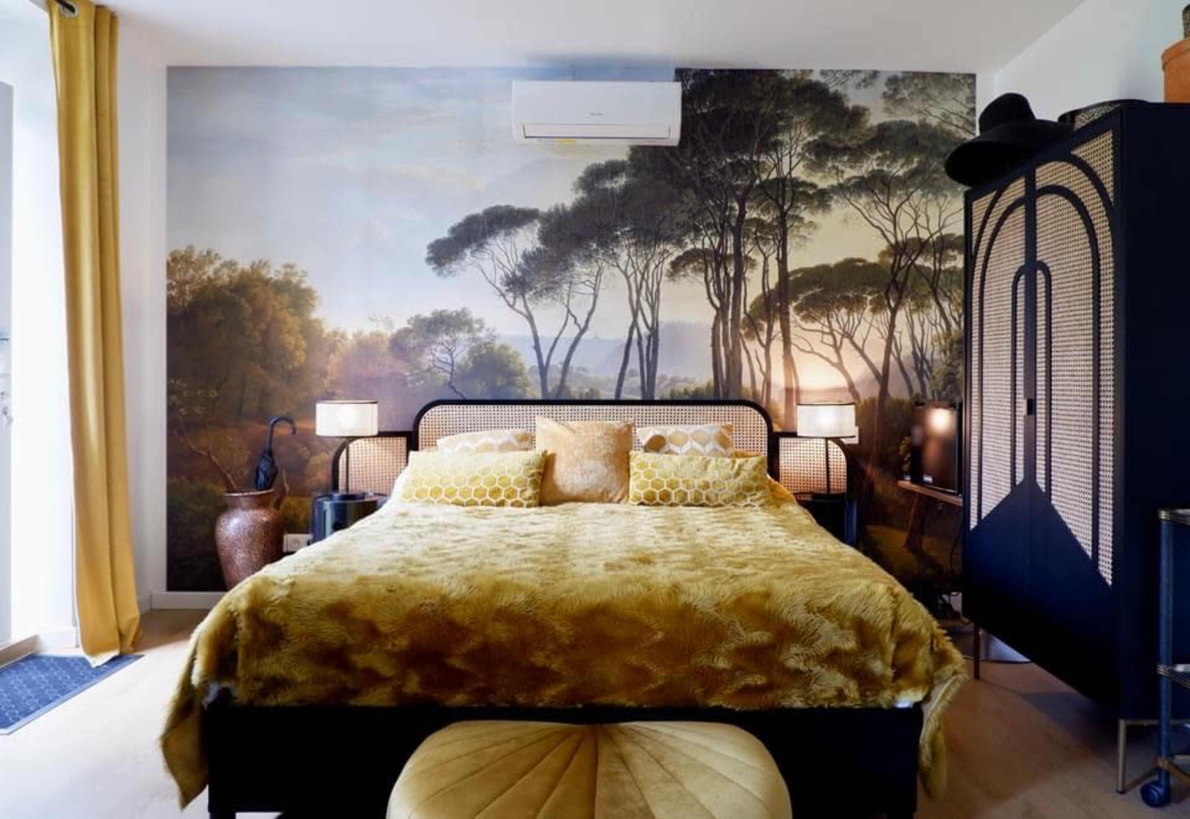 The image depicts a bedroom featuring a large bed with yellow bedding, framed by a mural of trees and complemented by stylish lamps and a wardrobe.