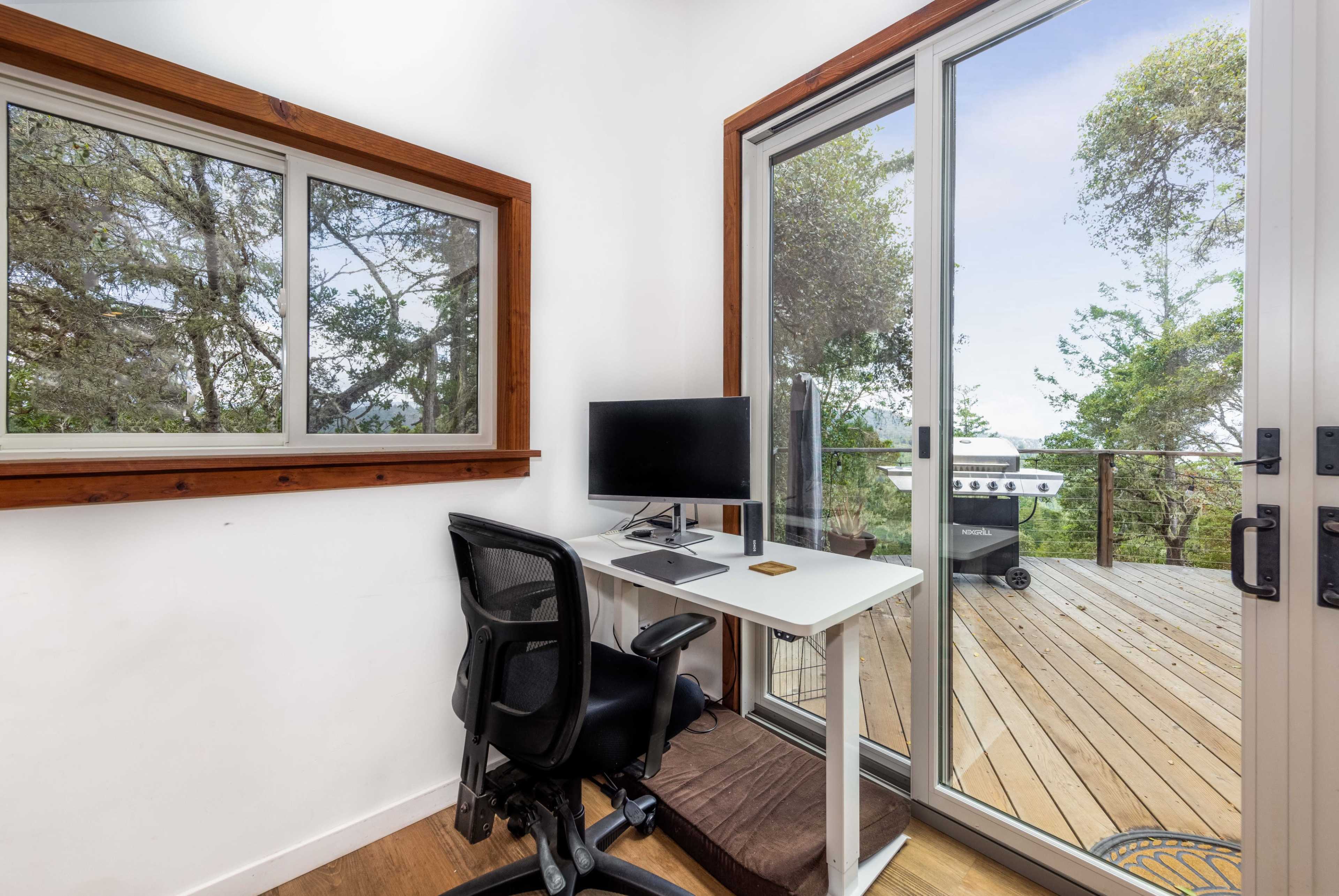 A small workspace with a desk and computer chair is positioned beside a large window and a sliding glass door that opens to a wooden deck.