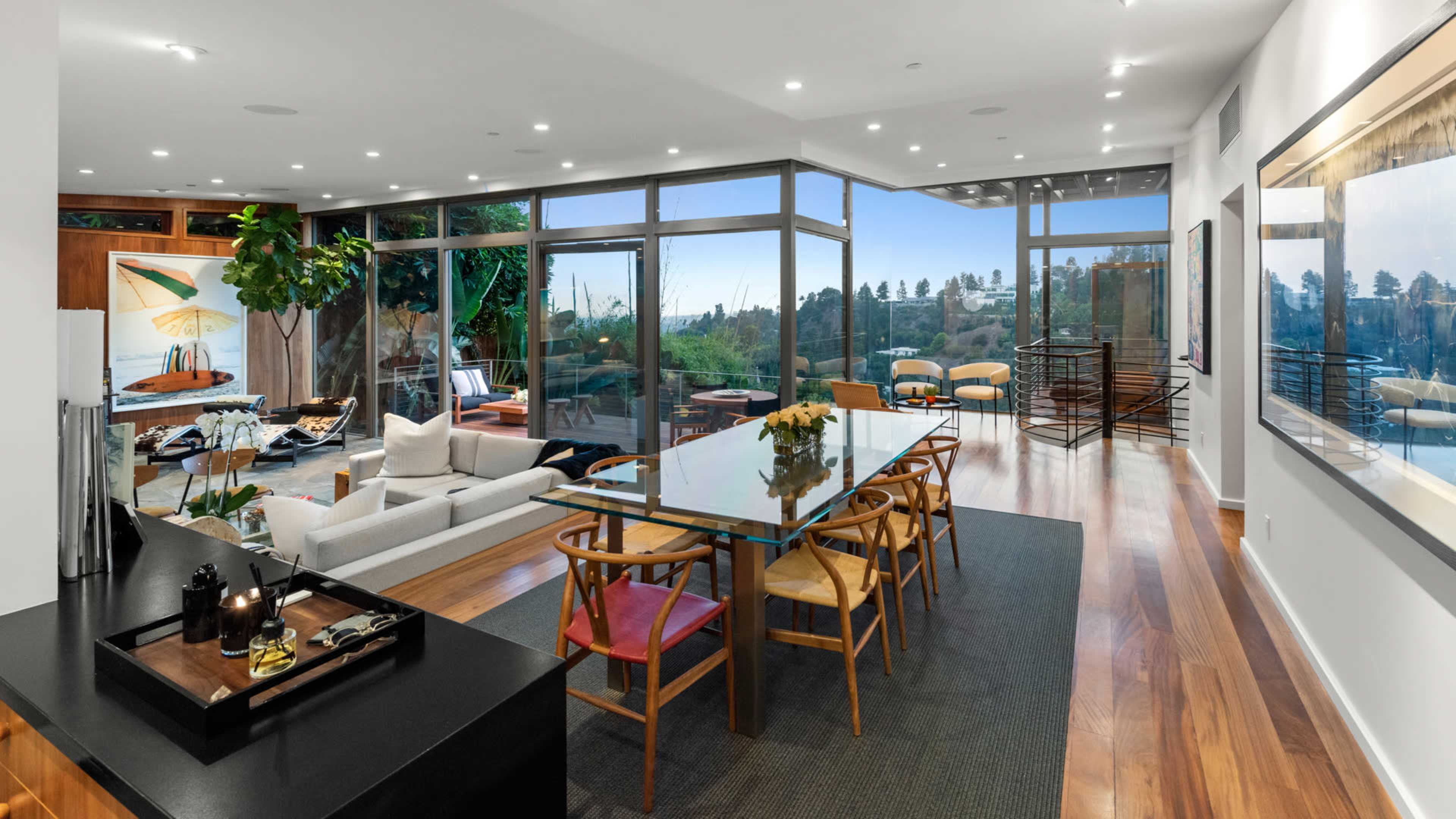 The image shows a modern, open-concept living and dining area with large windows that provide a view of a landscape.