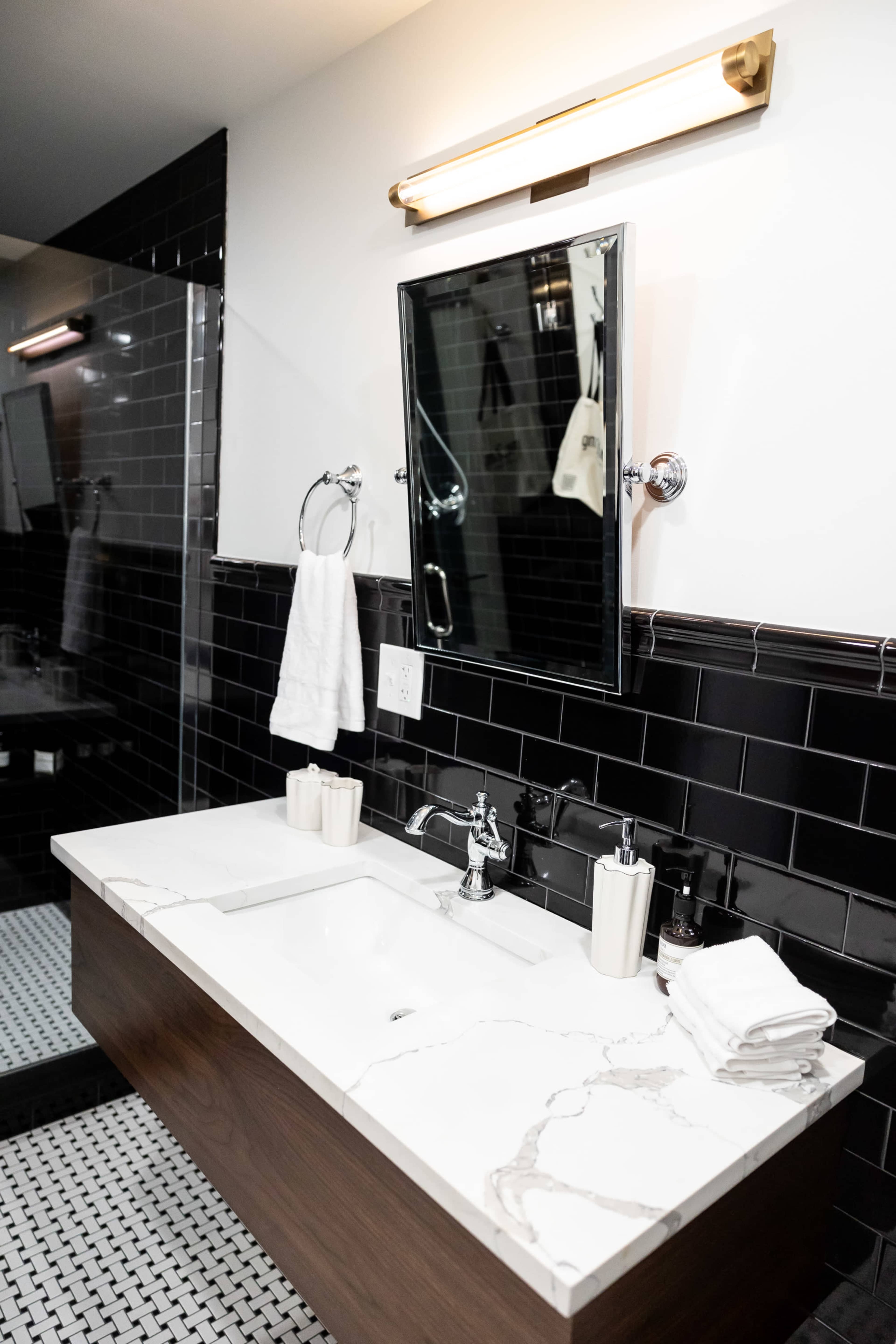 The image shows a modern bathroom featuring a wooden vanity with a white marble countertop, a mirror above, and black tiled walls.