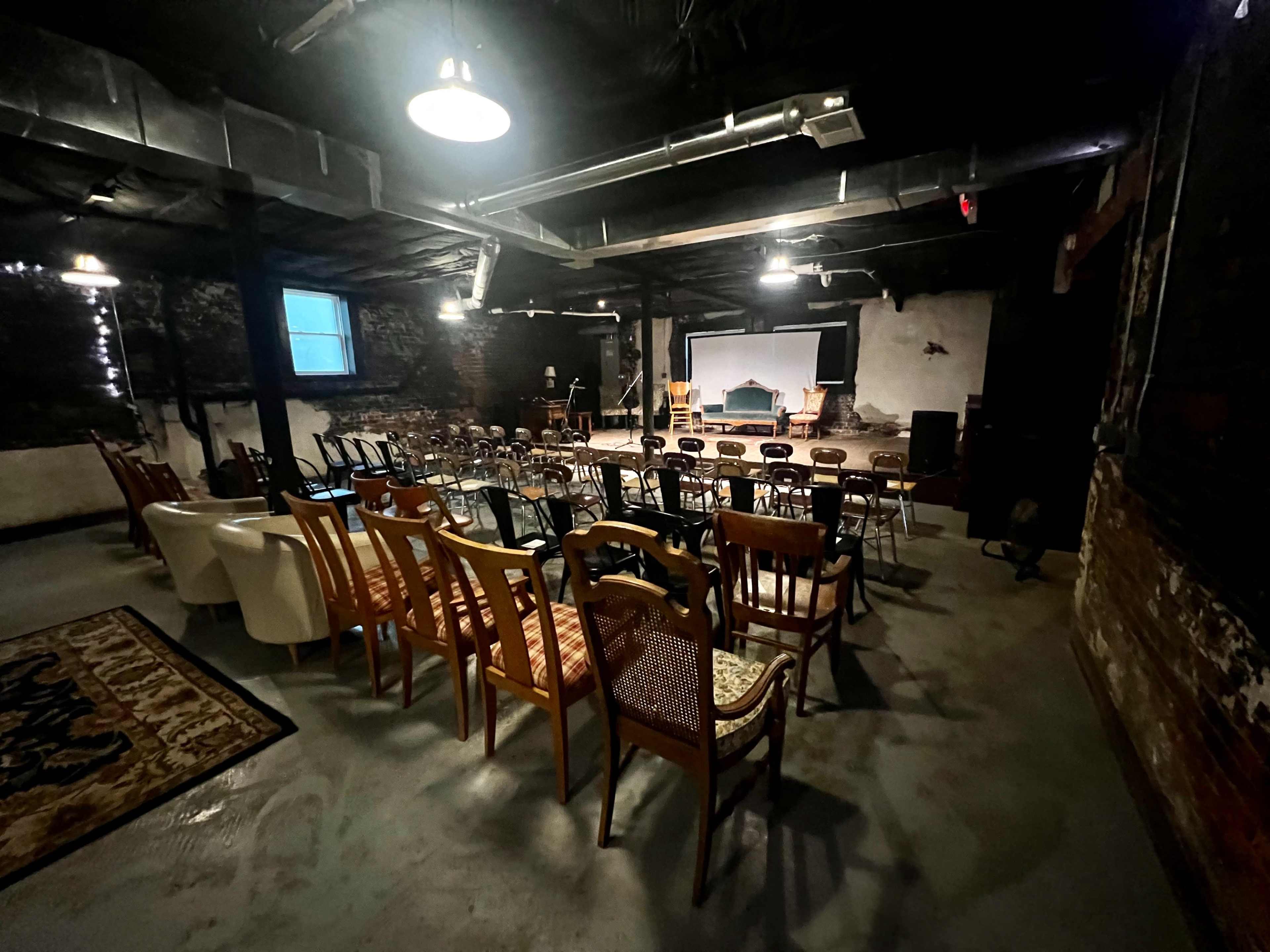 The image shows a dimly lit basement venue with rows of chairs arranged for an event, featuring a large screen at the front and a stage area.