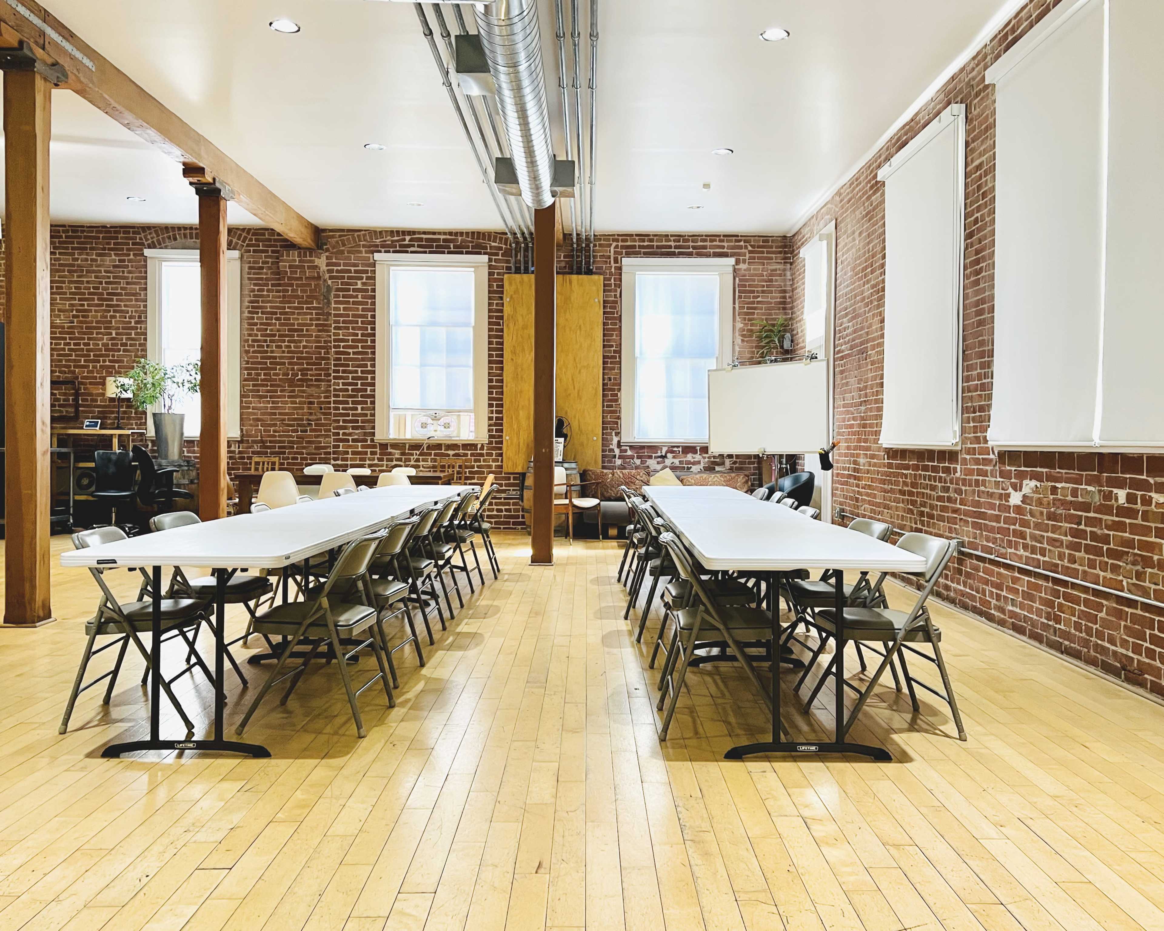 The image shows a spacious meeting room with two rows of tables and folding chairs arranged on a wooden floor, surrounded by exposed brick walls and large windows.