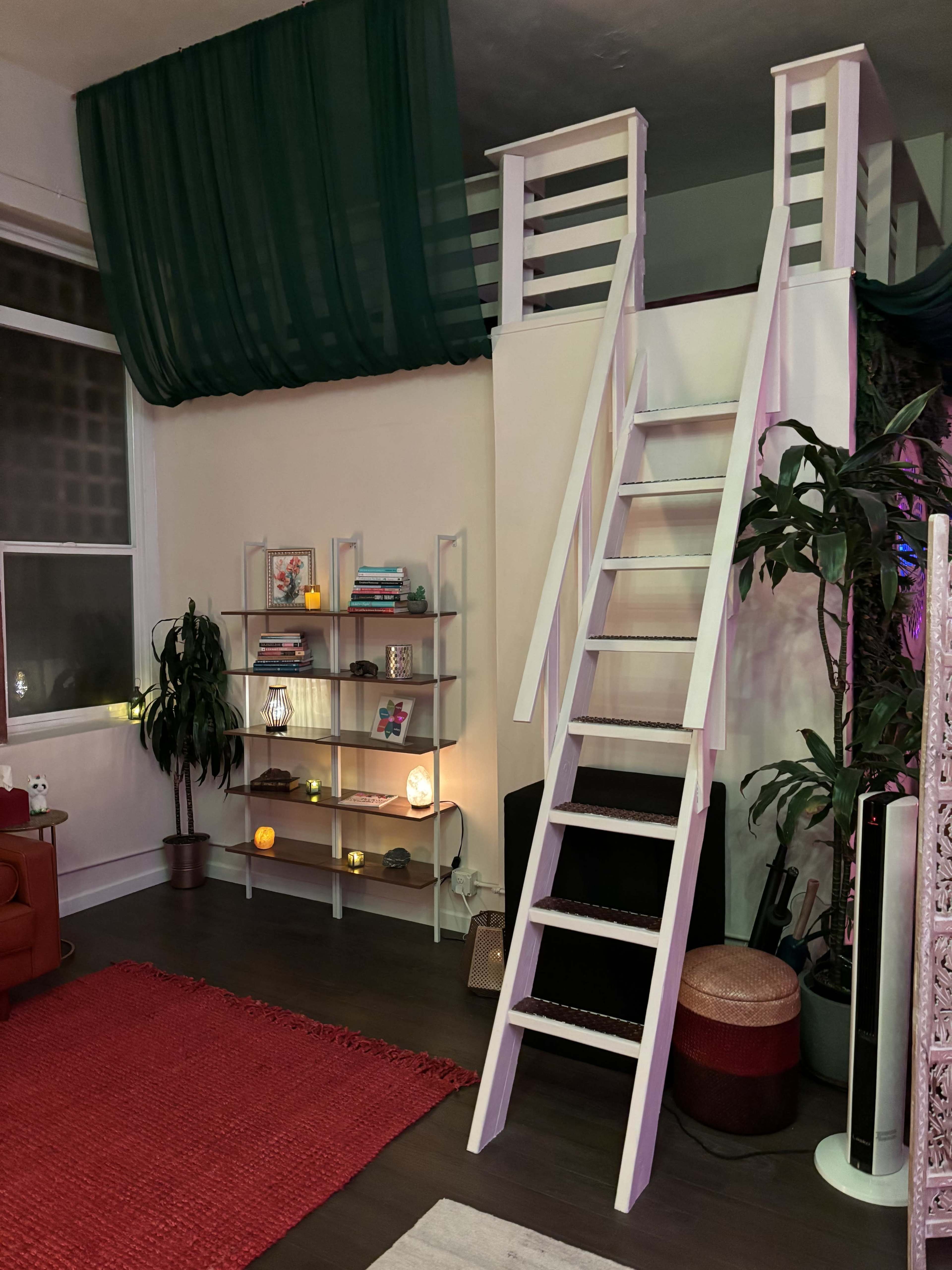 A cozy living space featuring a staircase leading to a loft area, with a shelf filled with books and decorative items, a red rug, and plants.