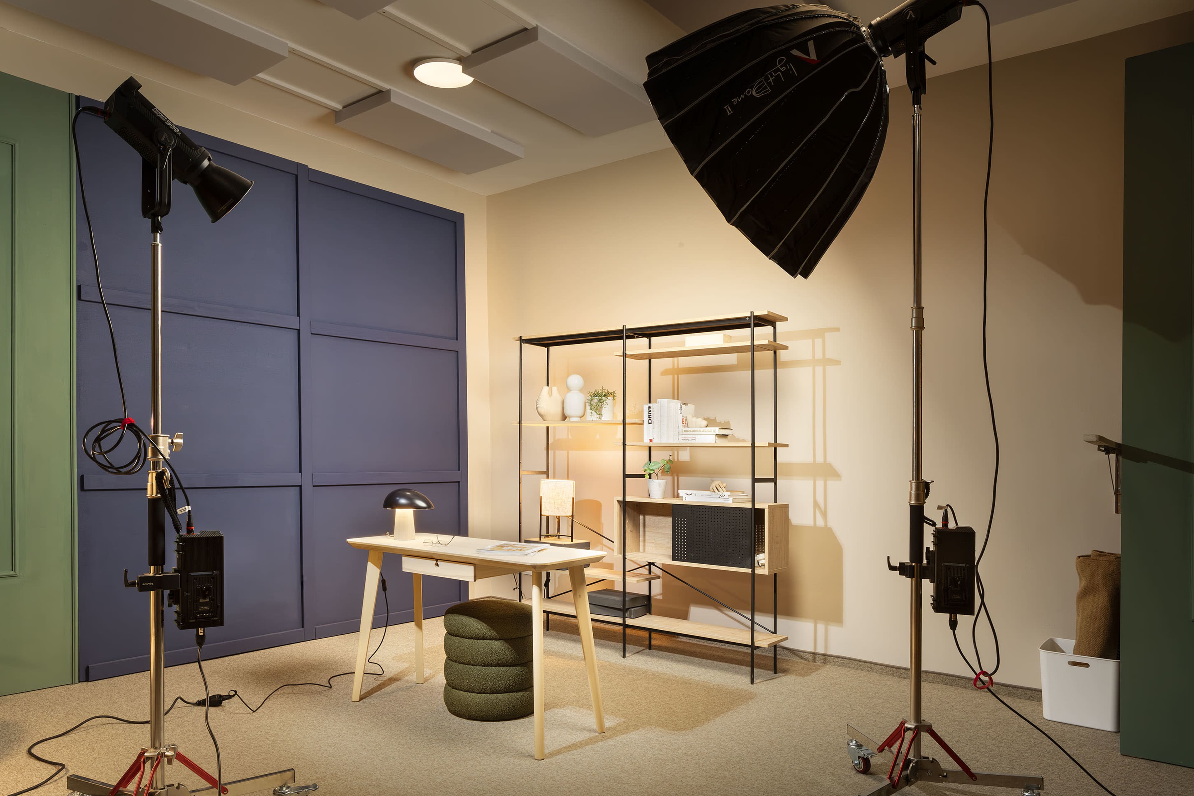 A modern studio setup features a desk with a lamp and decorative items, surrounded by lighting equipment and shelving against a backdrop of colored walls.