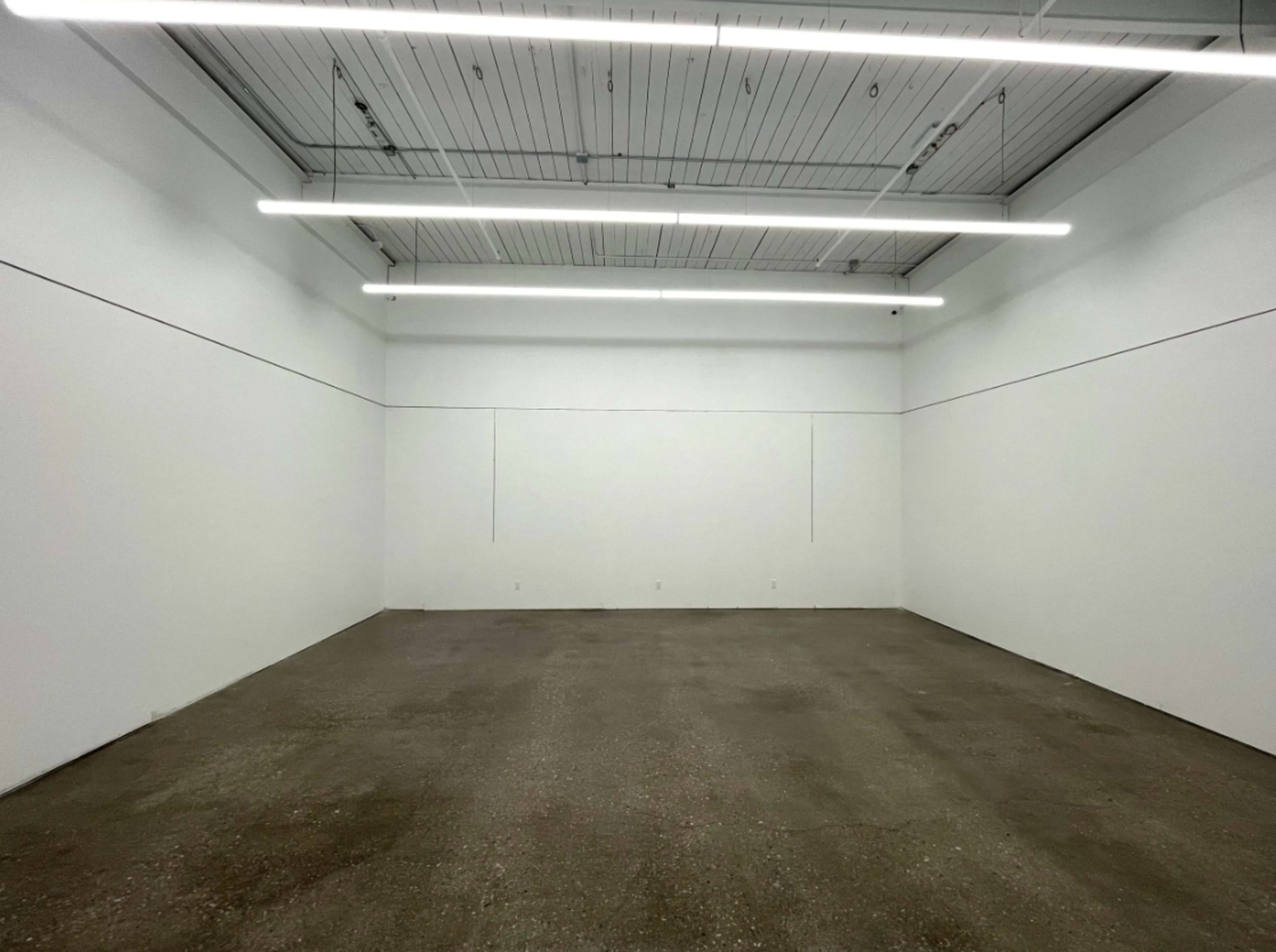 The image shows an empty room with plain white walls, concrete flooring, and overhead fluorescent lighting.