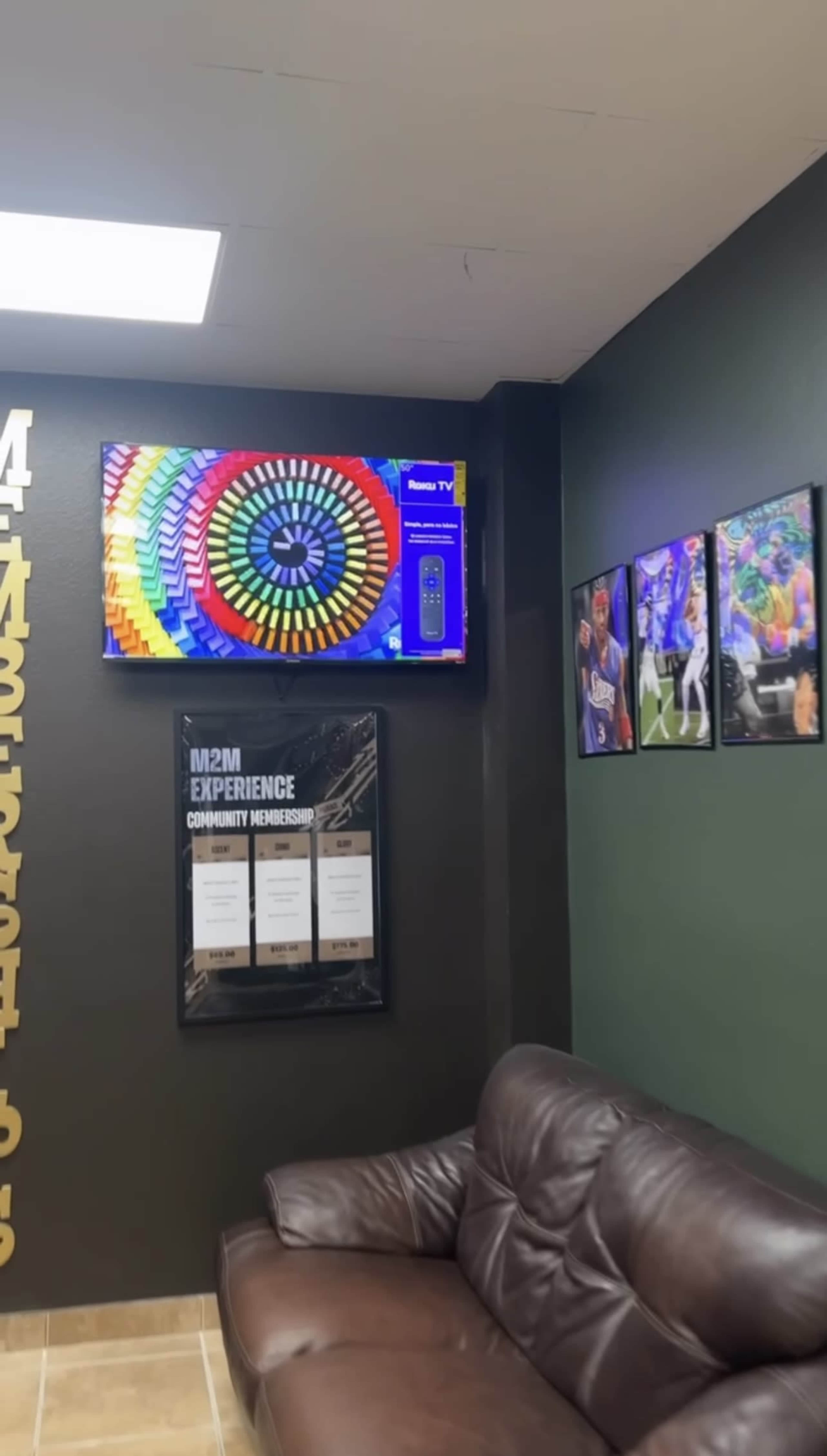 The image shows a lounge area featuring a brown couch, a television displaying a colorful circular graphic, and framed photos on the wall.