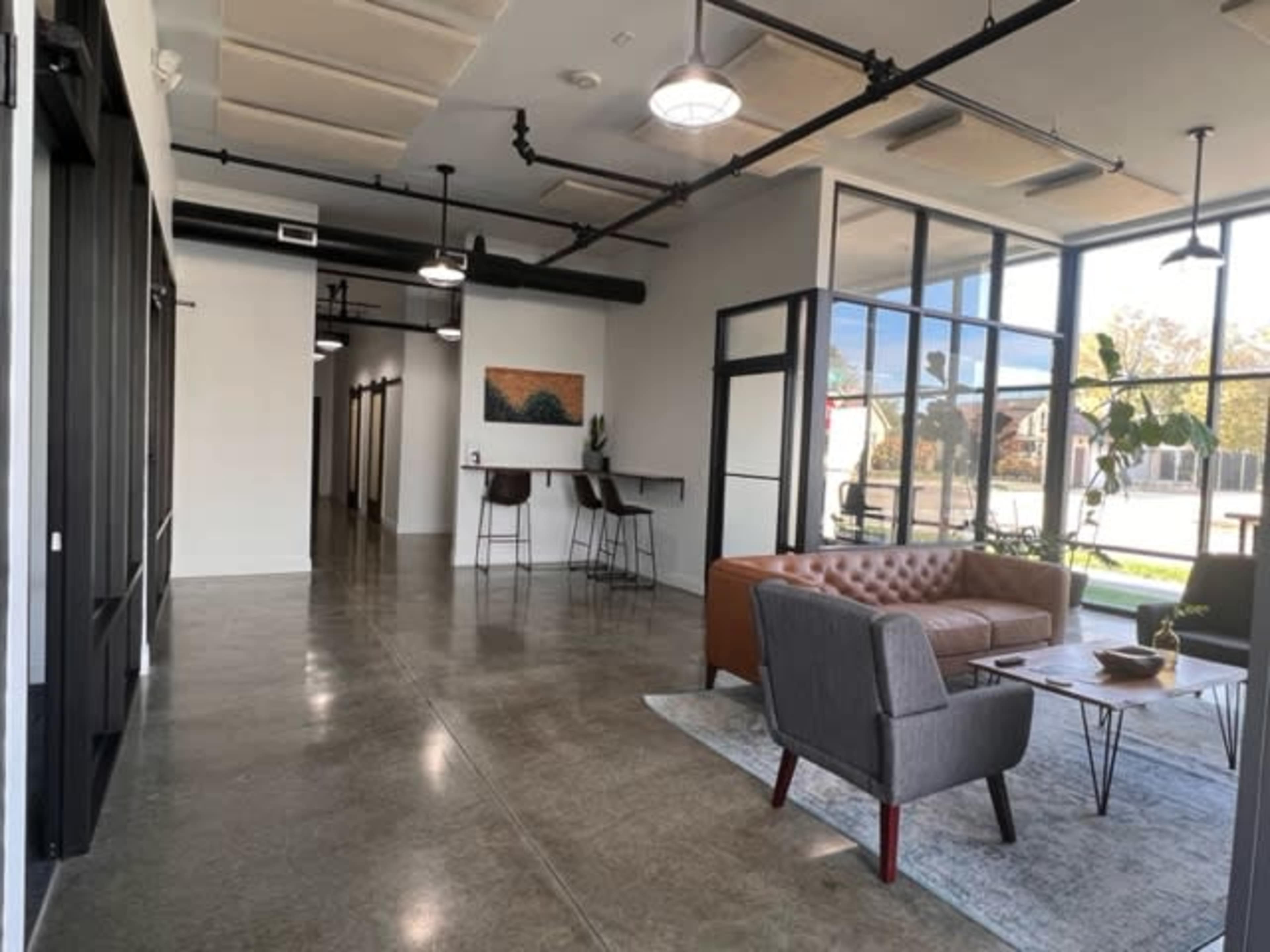 The image shows a modern open workspace with a lounge area featuring a brown leather sofa and a coffee table, alongside a bar with high chairs and large windows letting in natural light.