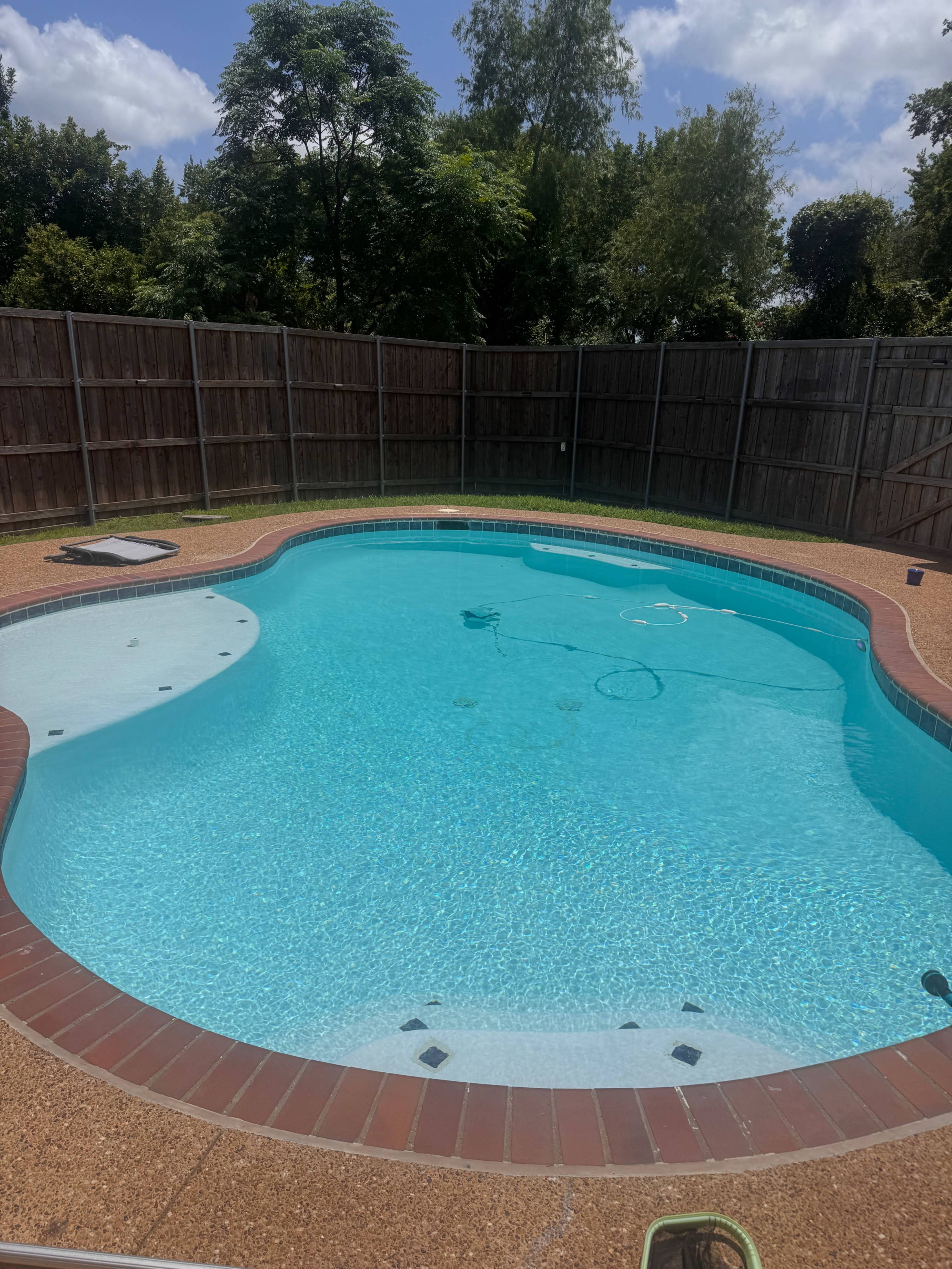 The image shows a clear swimming pool surrounded by a wooden fence and green trees in the background.