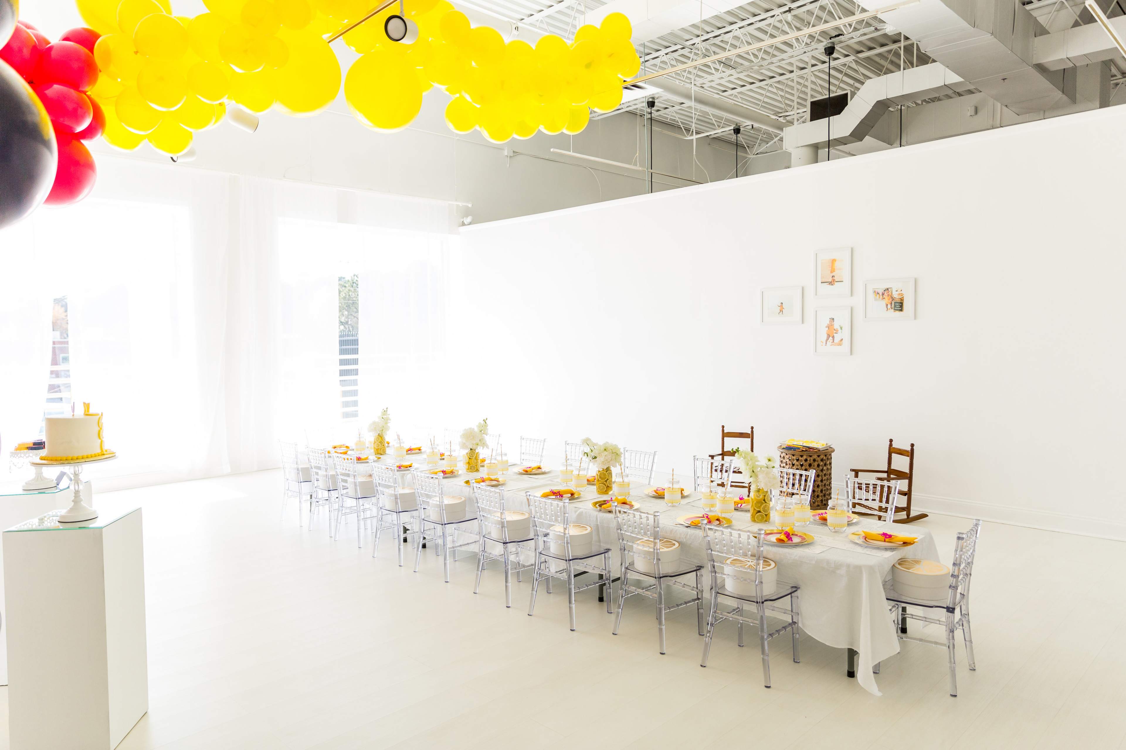 A long, elegantly set dining table with white linens and yellow decorations is arranged in a bright, airy room featuring large windows.