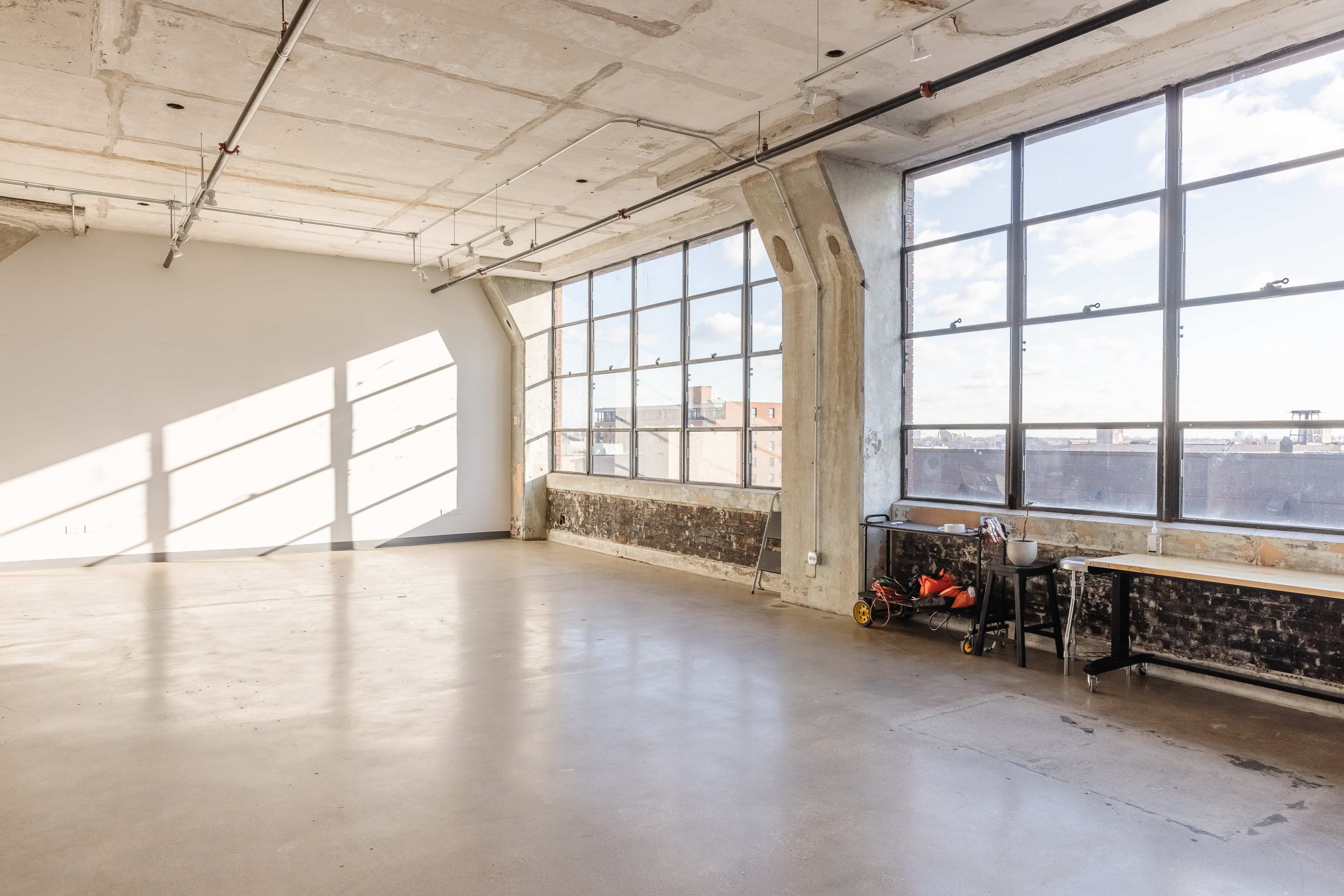 A spacious room with large windows allowing natural light to fill the concrete-floored area, featuring a simple table and a few items along one wall.