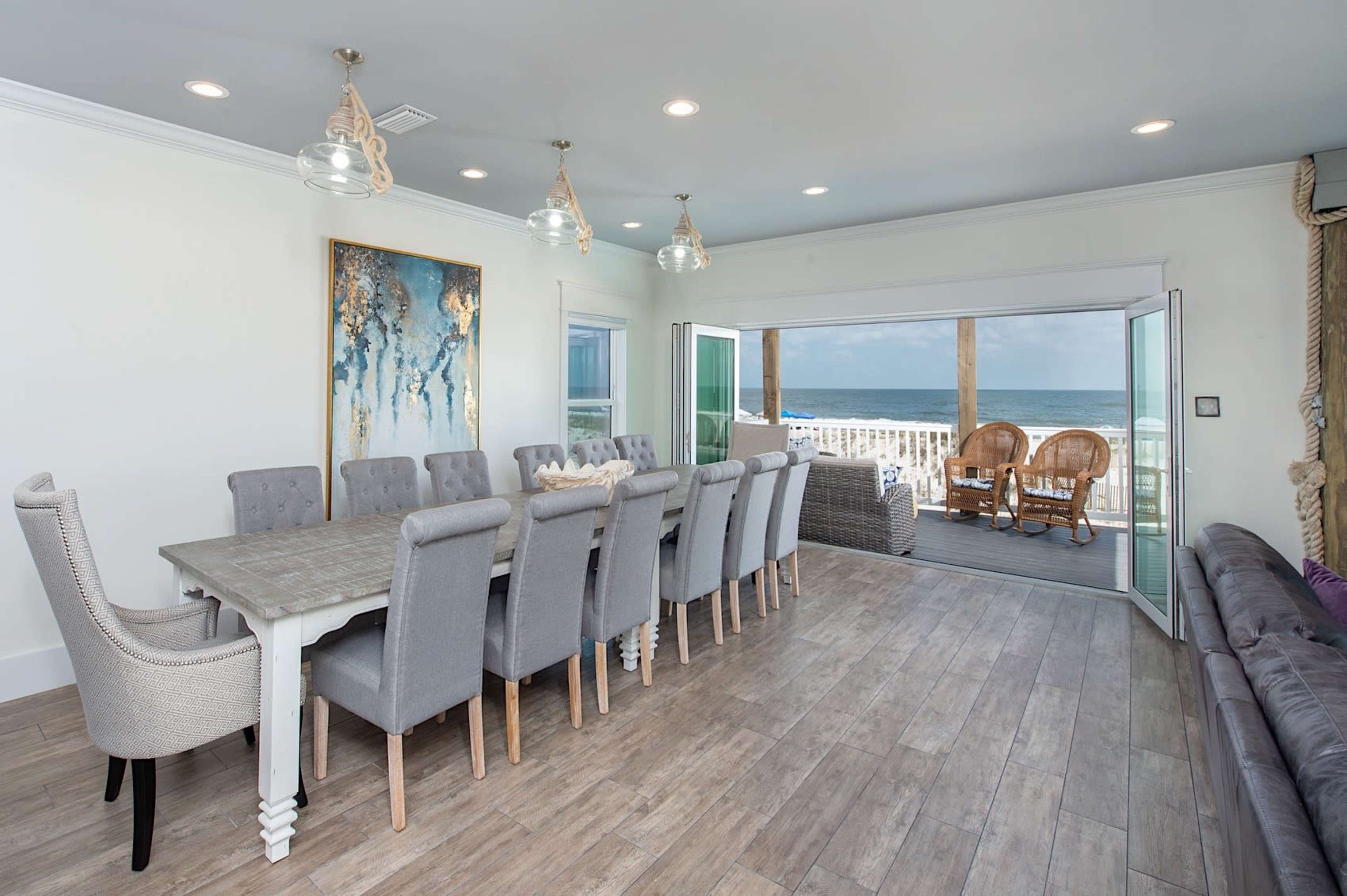 A spacious dining room with a long table surrounded by upholstered chairs opens to a deck overlooking the ocean.