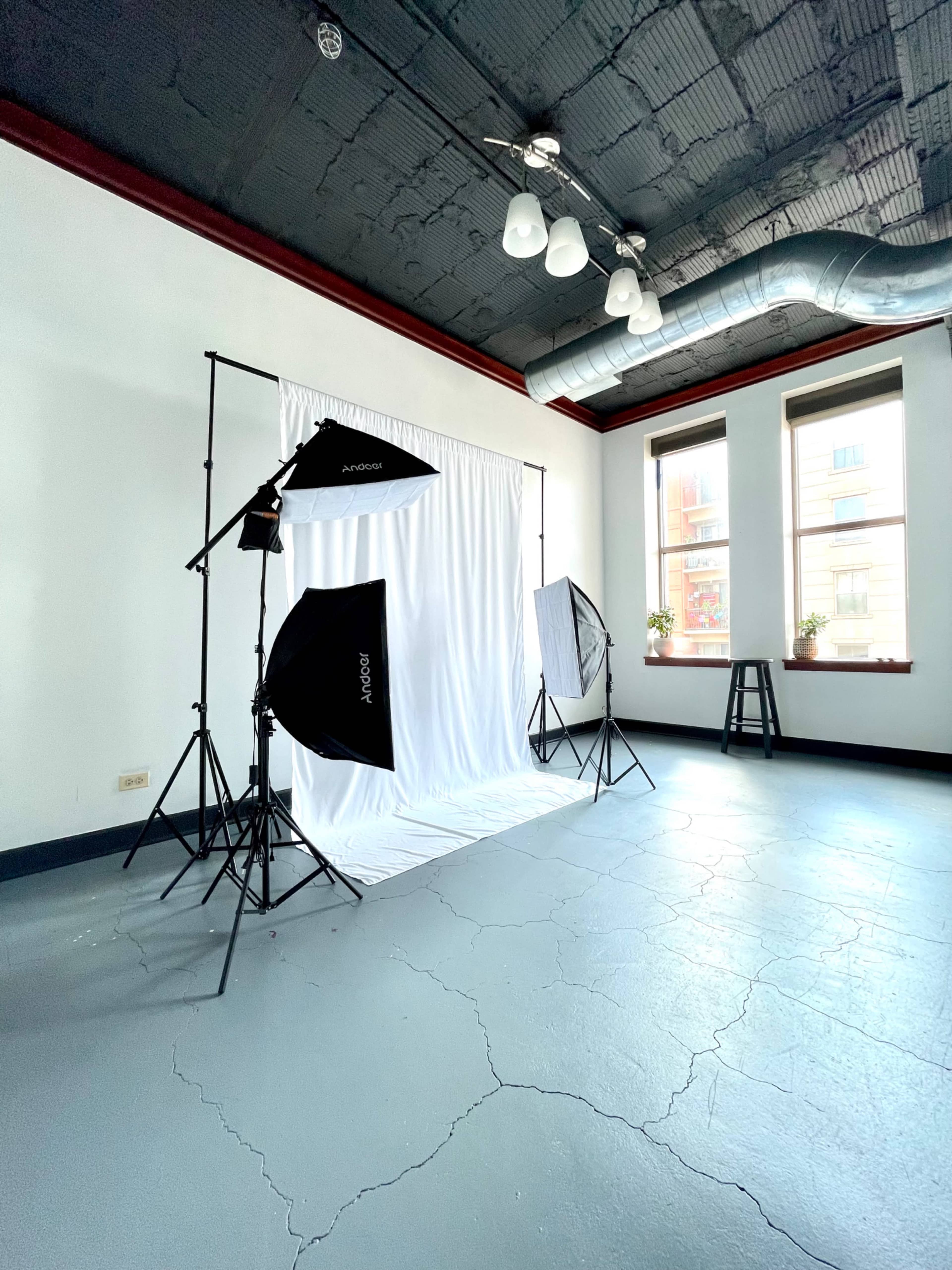 A photography studio with a white backdrop, softbox lights on stands, and a wooden stool in a brightly lit room.