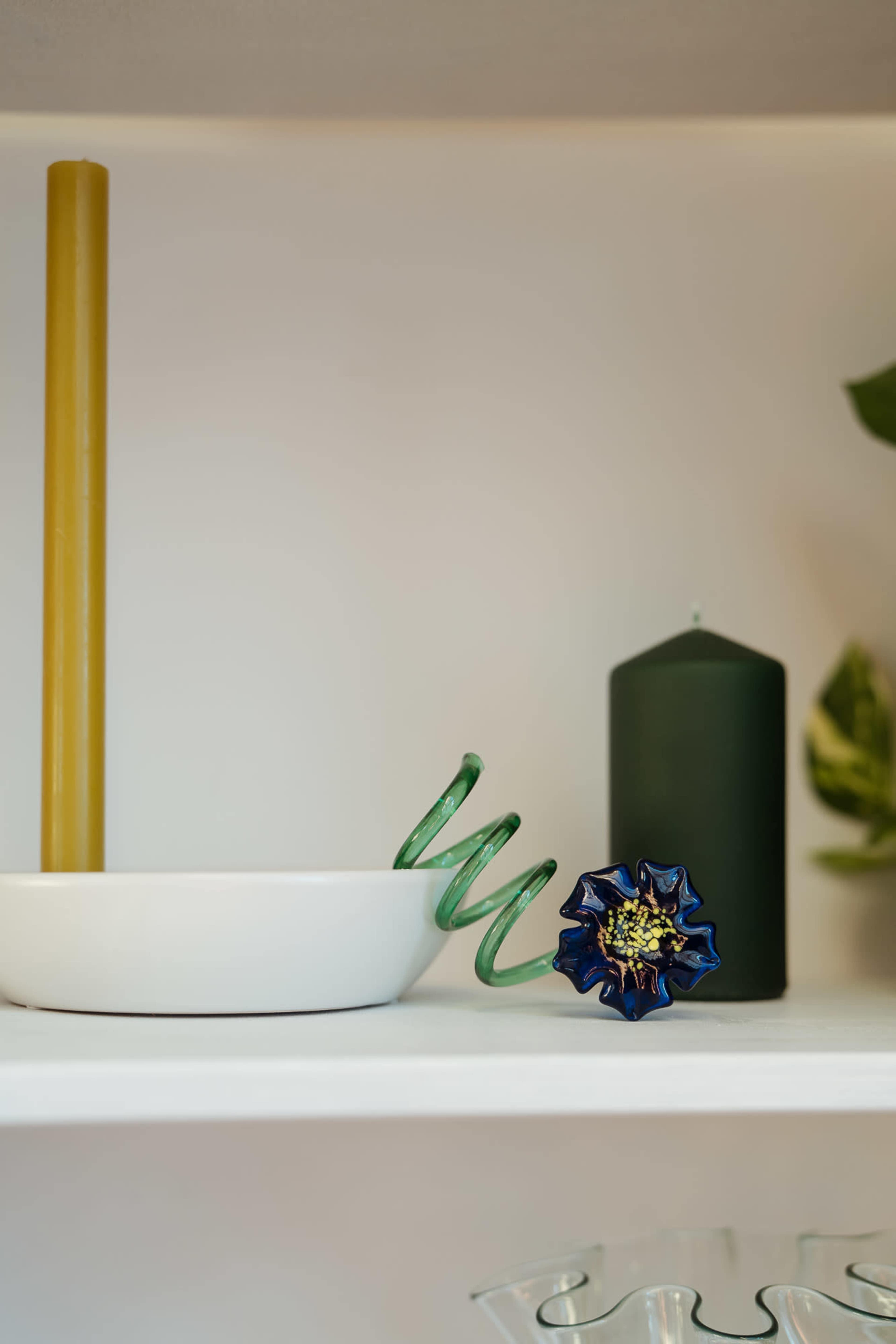 The image features a white dish with a green spiral object, a yellow candle, a dark green candle, and a blue flower-shaped decorative item on a shelf.
