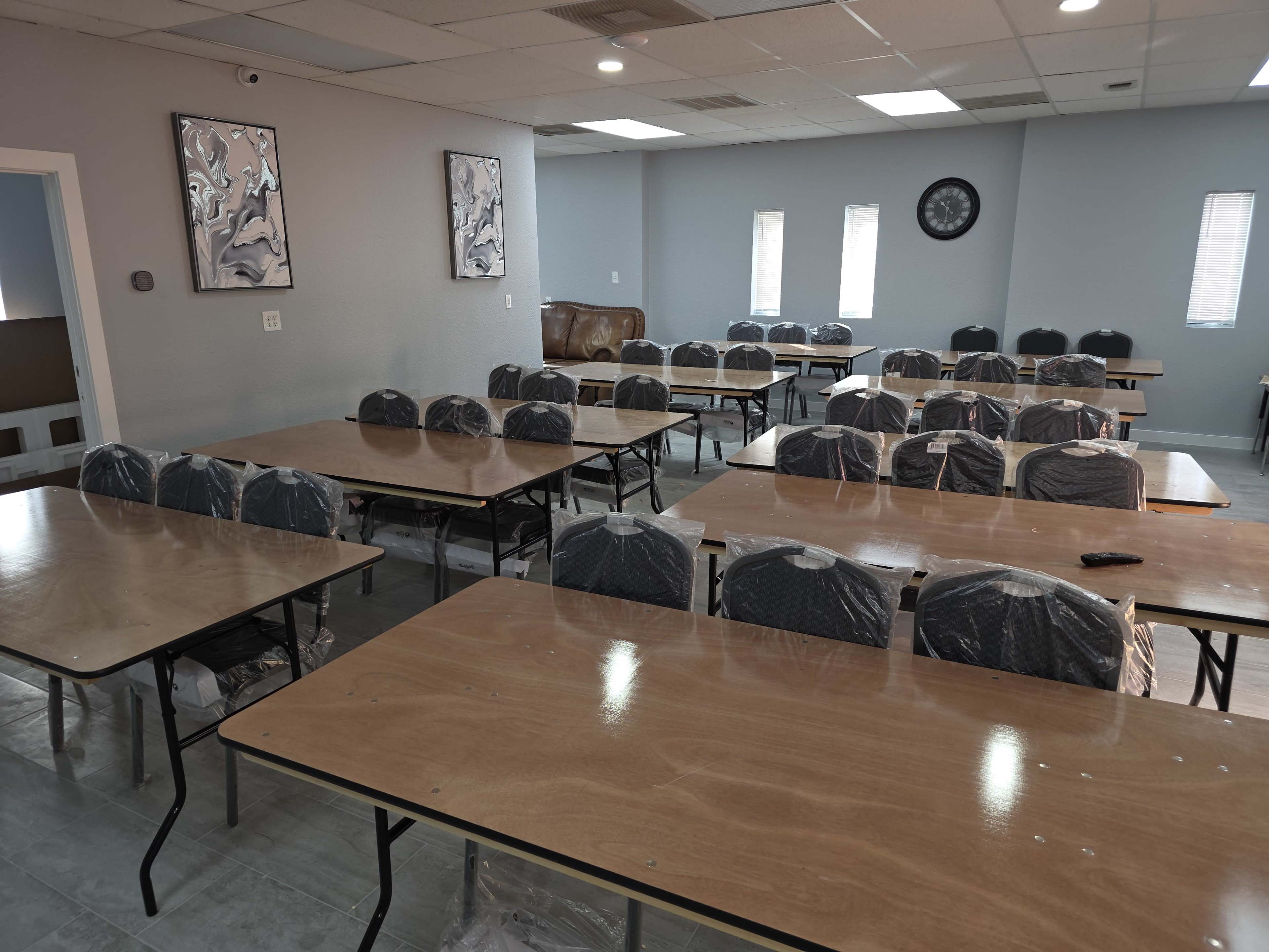 The image shows a room set up with multiple tables and chairs, with each chair covered in plastic, indicating preparation for an event or meeting.