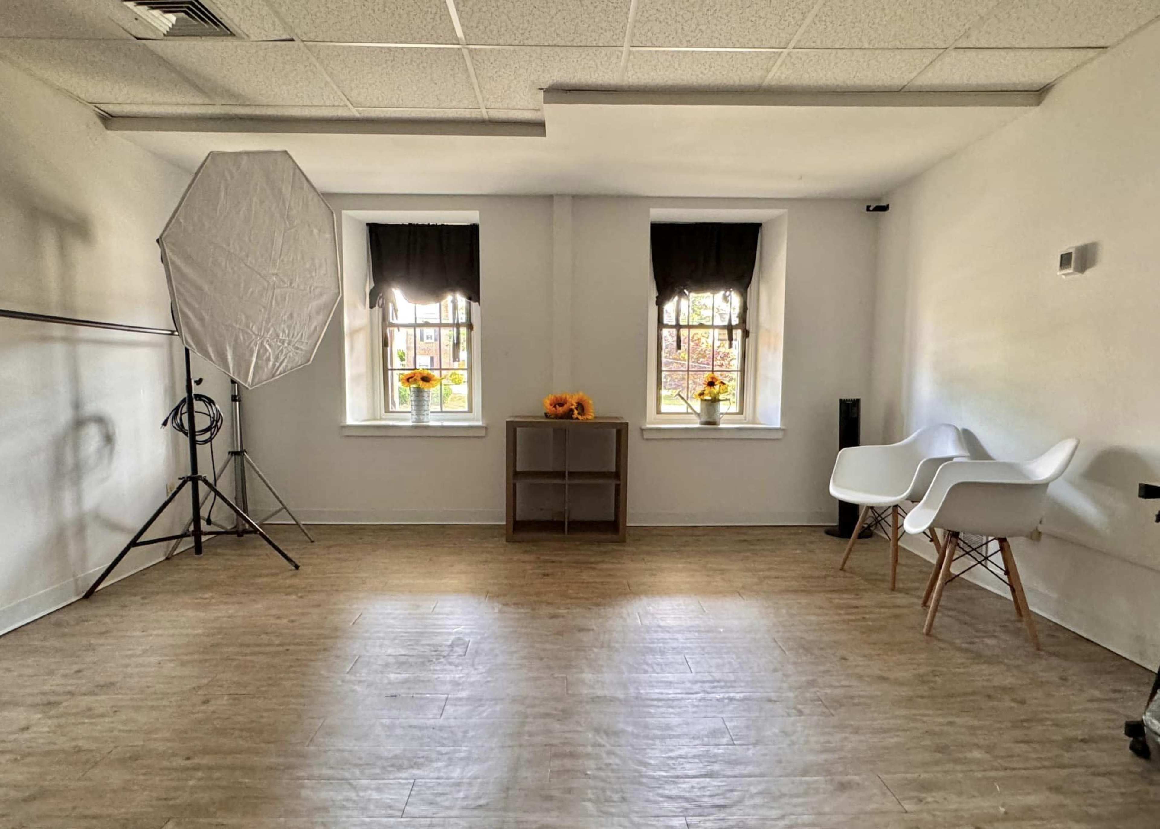 The image shows a well-lit room with two windows, a photography backdrop, a small wooden shelf, and minimalist white chairs.