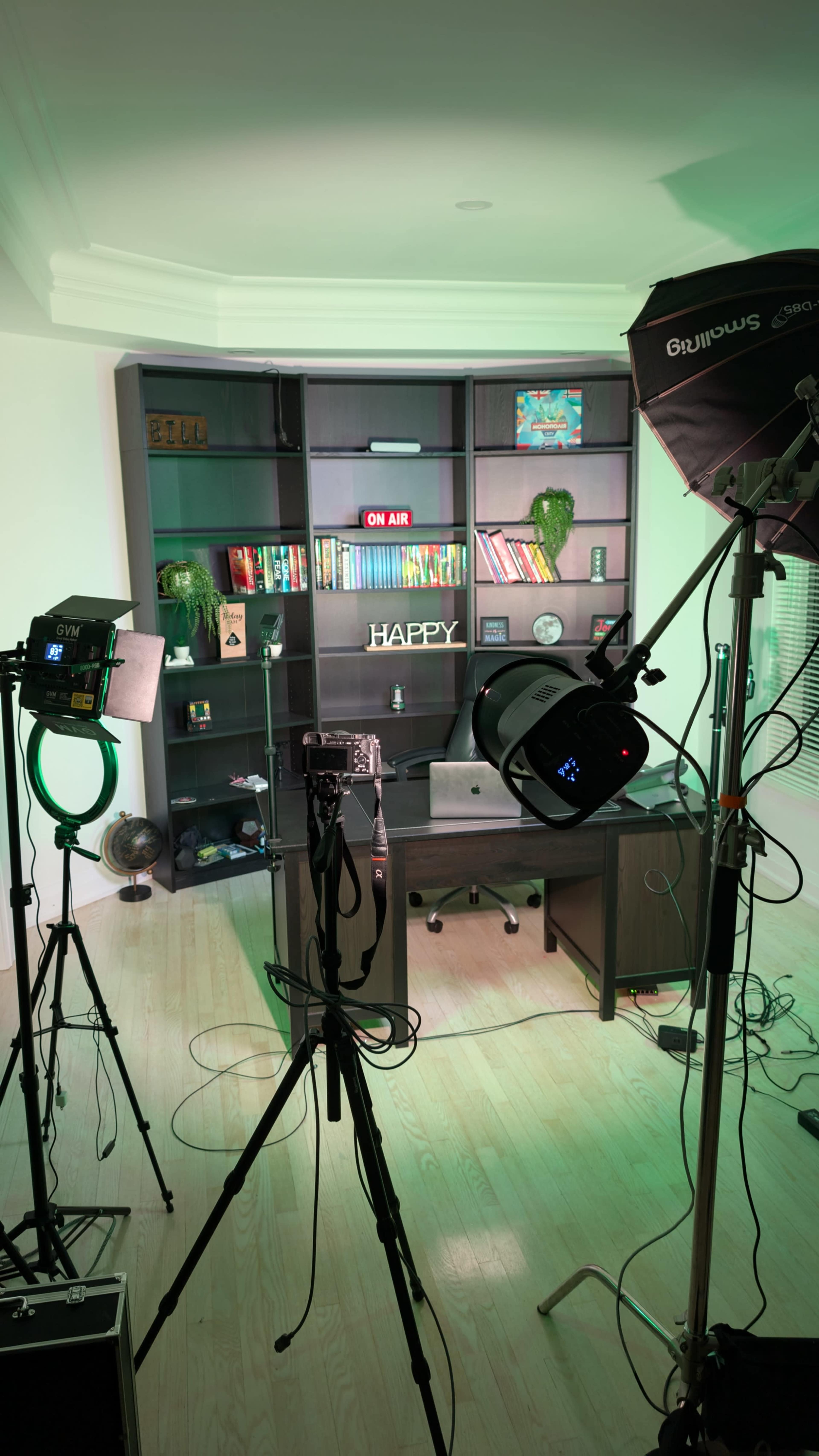 The image shows a modern recording studio setup with a desk, bookshelves filled with books, and lighting equipment positioned around the room.
