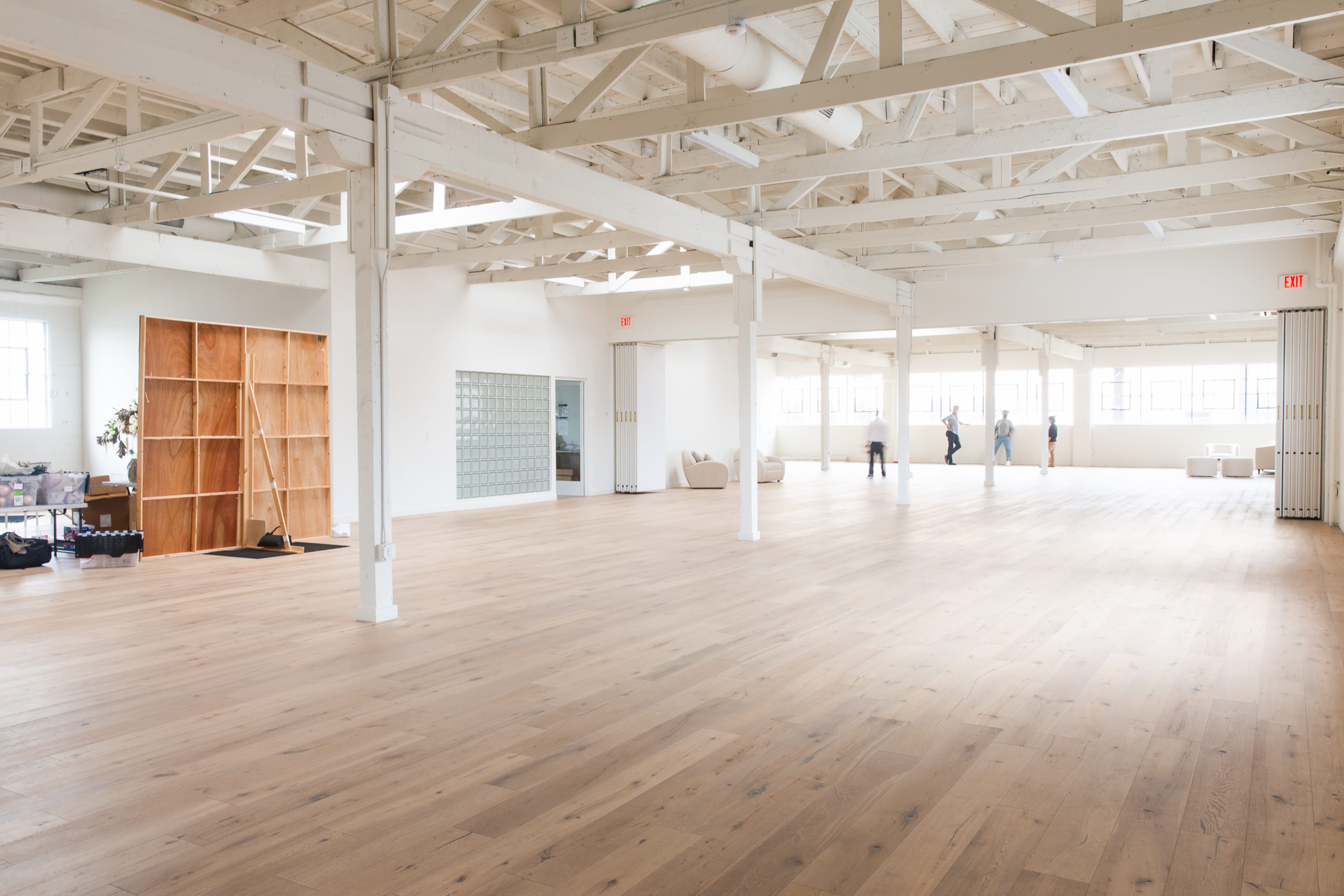 The image shows a spacious, empty room with wooden floors, large windows, and exposed ceiling beams.