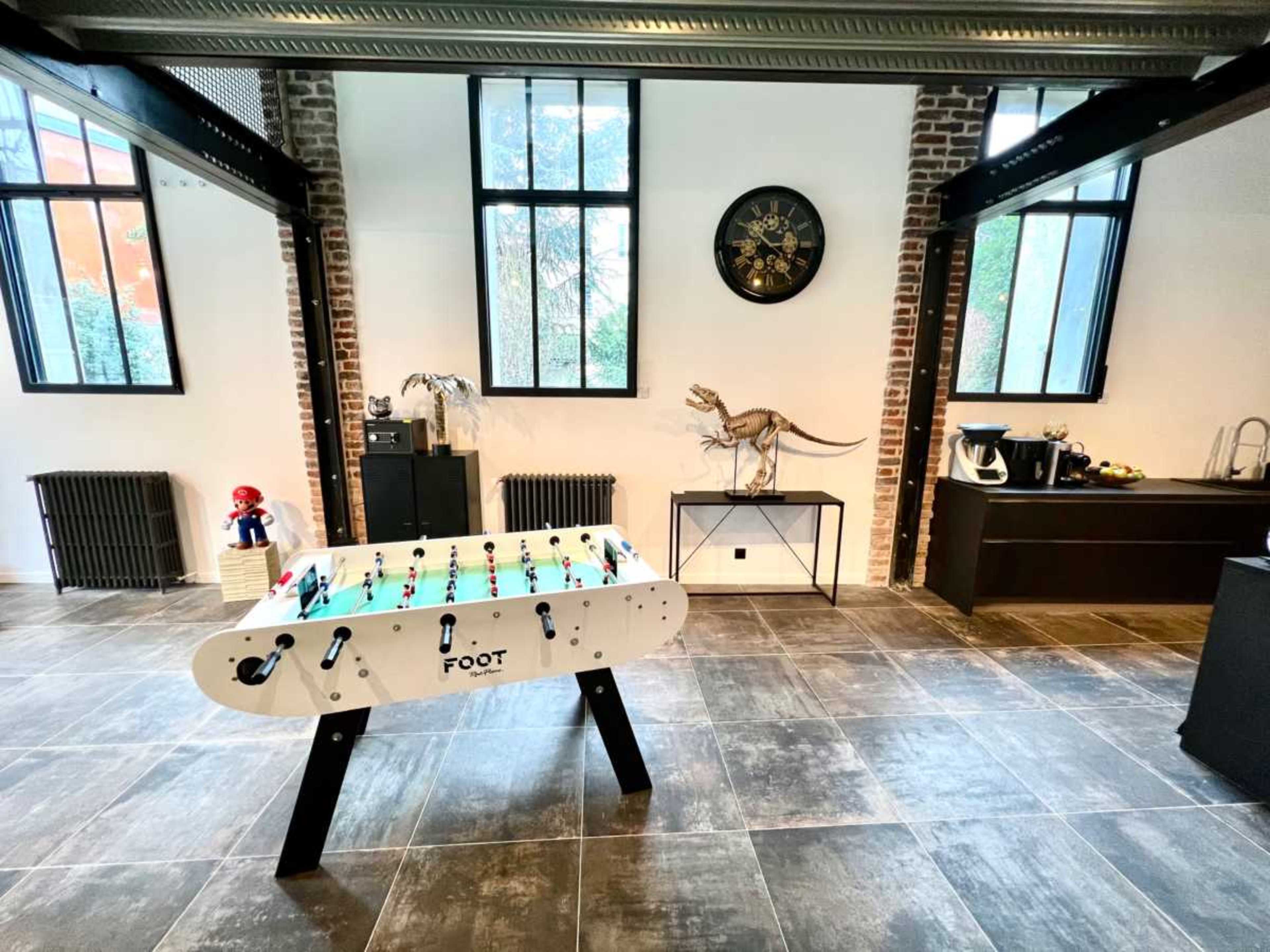 A spacious room with a foosball table in the center, large windows, and various decorative elements, including a dinosaur figurine and a modern sideboard.