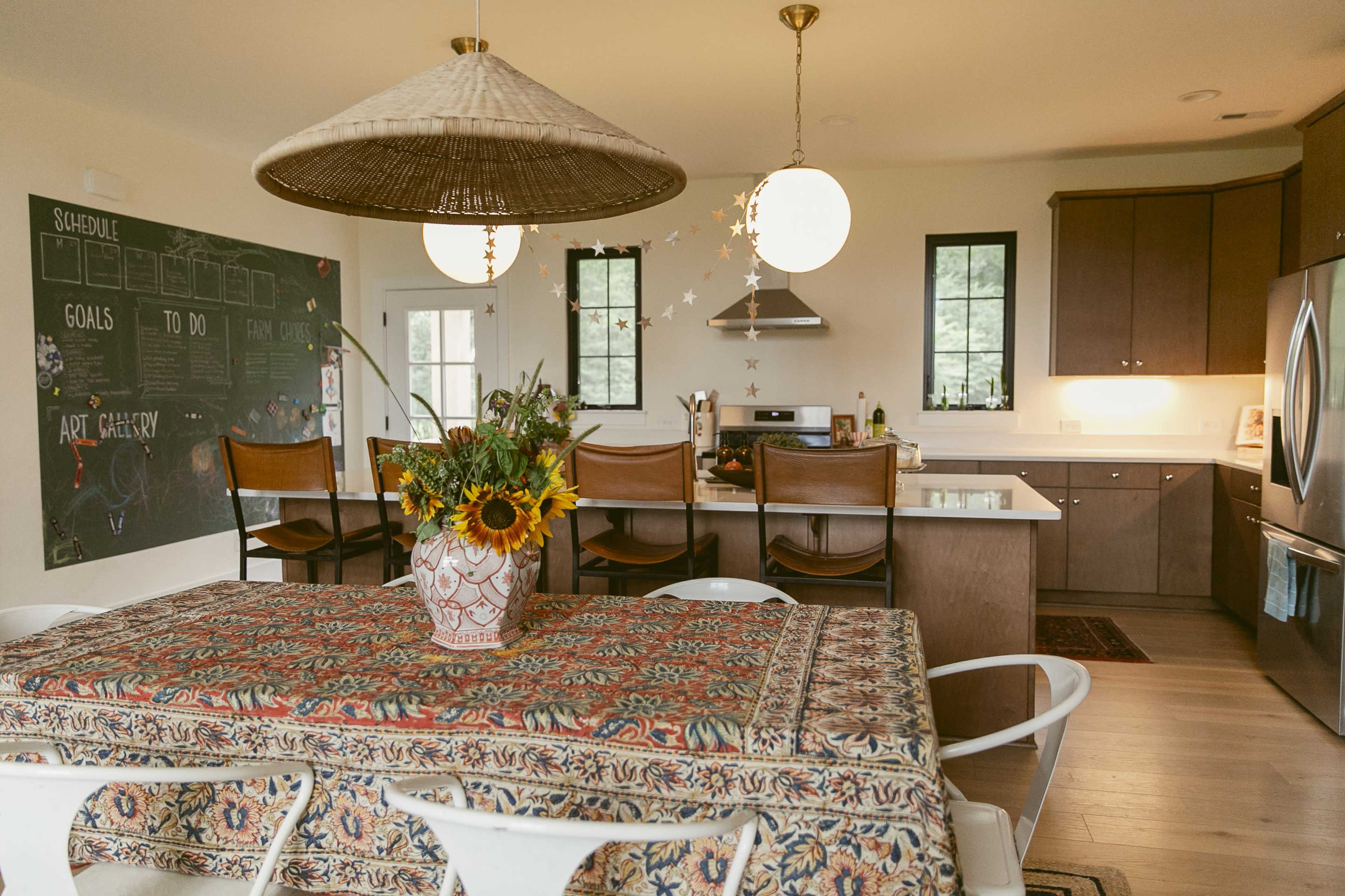 The image shows a modern kitchen with a dining table adorned with a patterned tablecloth, surrounded by white chairs, and features a chalkboard wall, wooden cabinets, and pendant lighting.