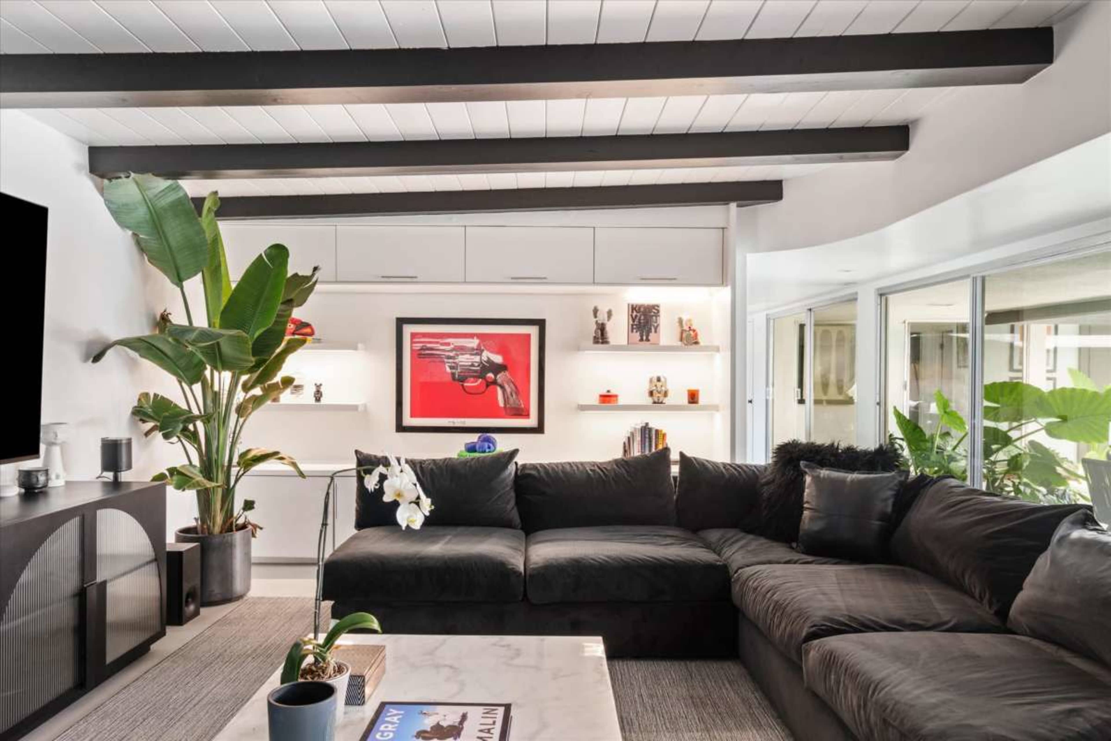 The image shows a modern living room featuring a black sectional sofa, a coffee table, and a white feature wall with decorative shelves and a striking artwork of a gun.