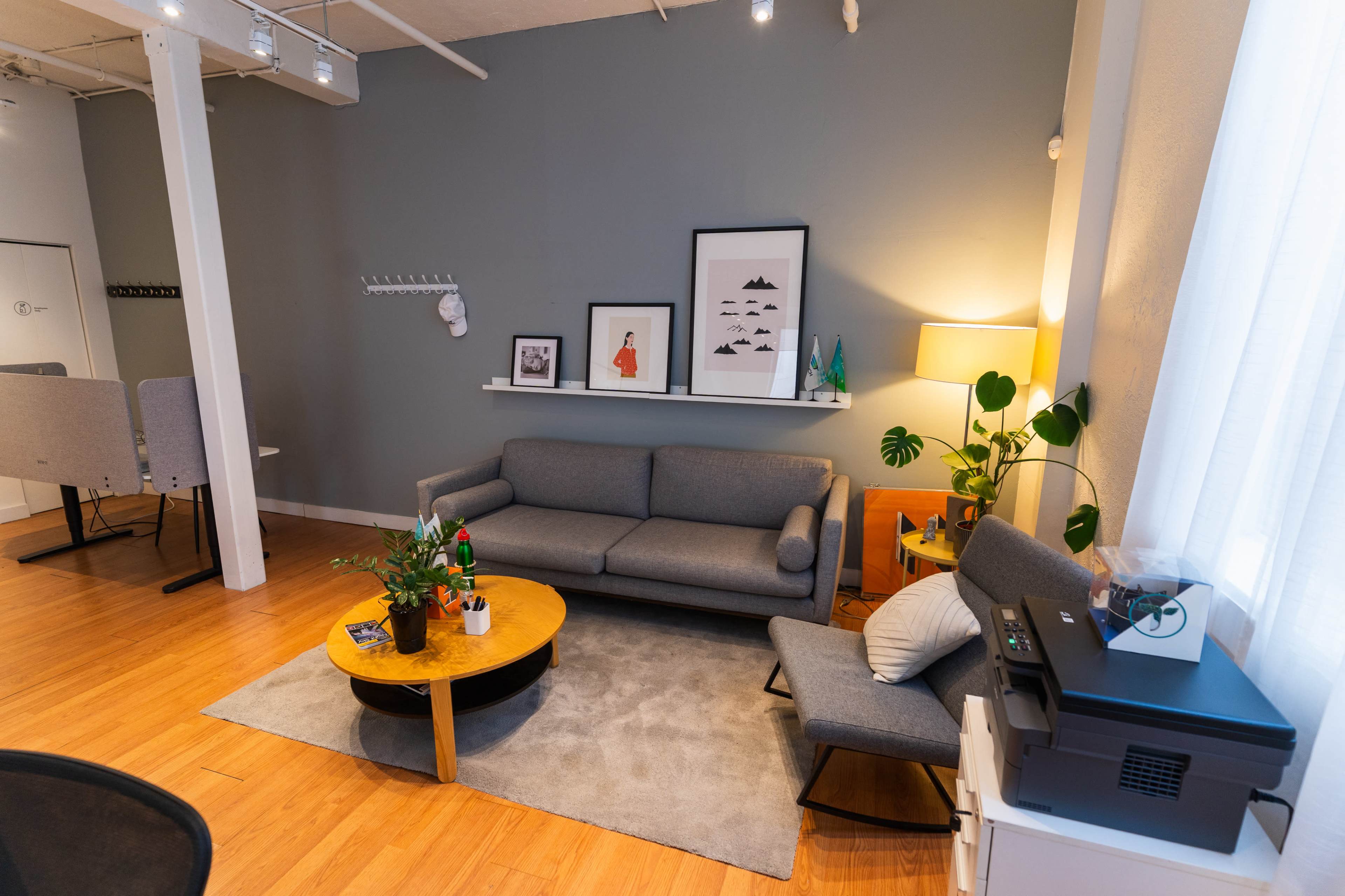 The image shows a modern interior living space featuring a gray sofa, a round wooden coffee table, a lamp, and decorative plants, with a light gray wall and large window in the background.