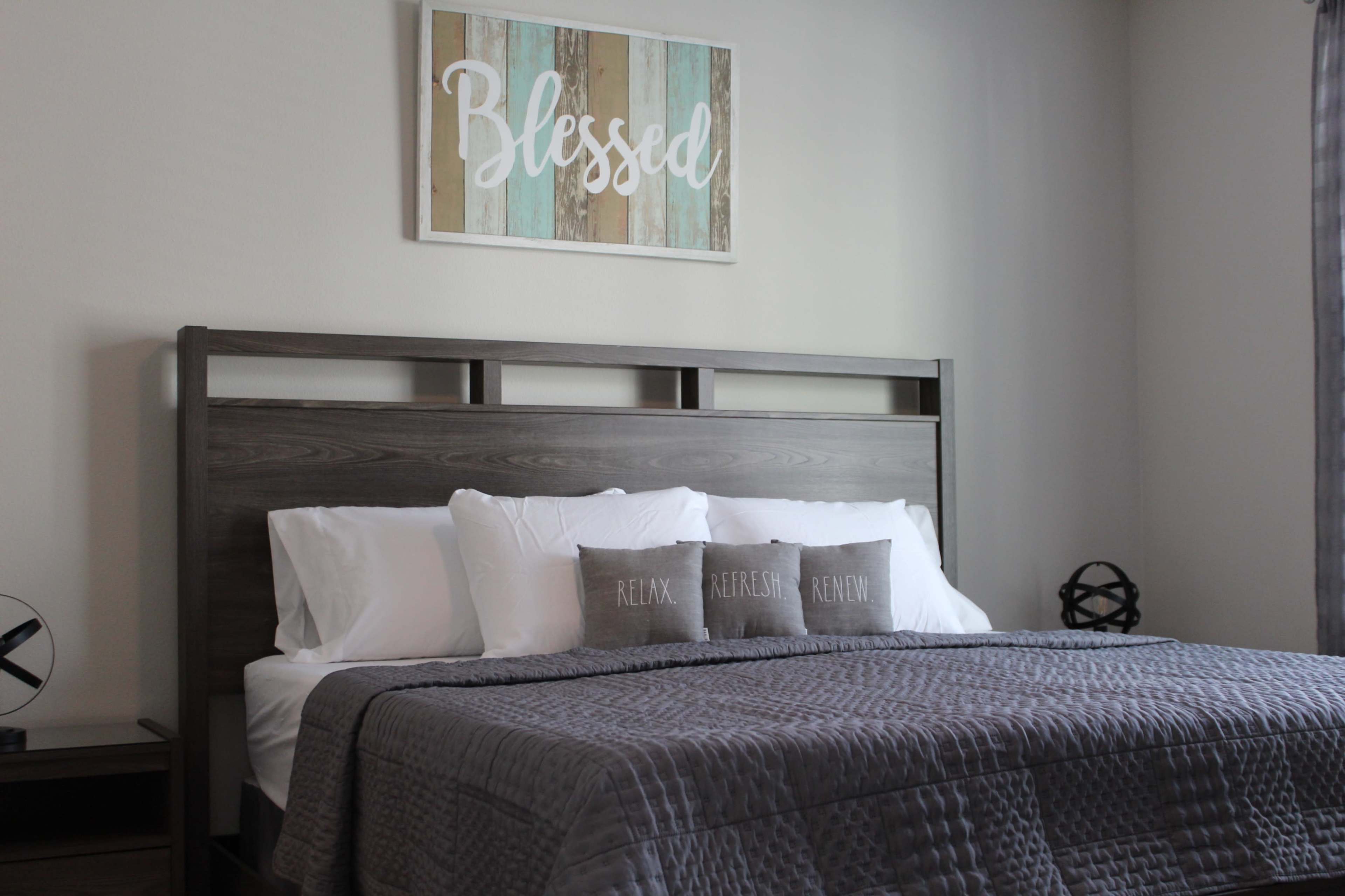 A neatly arranged bedroom features a wooden bed frame with a gray quilt and decorative pillows on a light-colored wall.