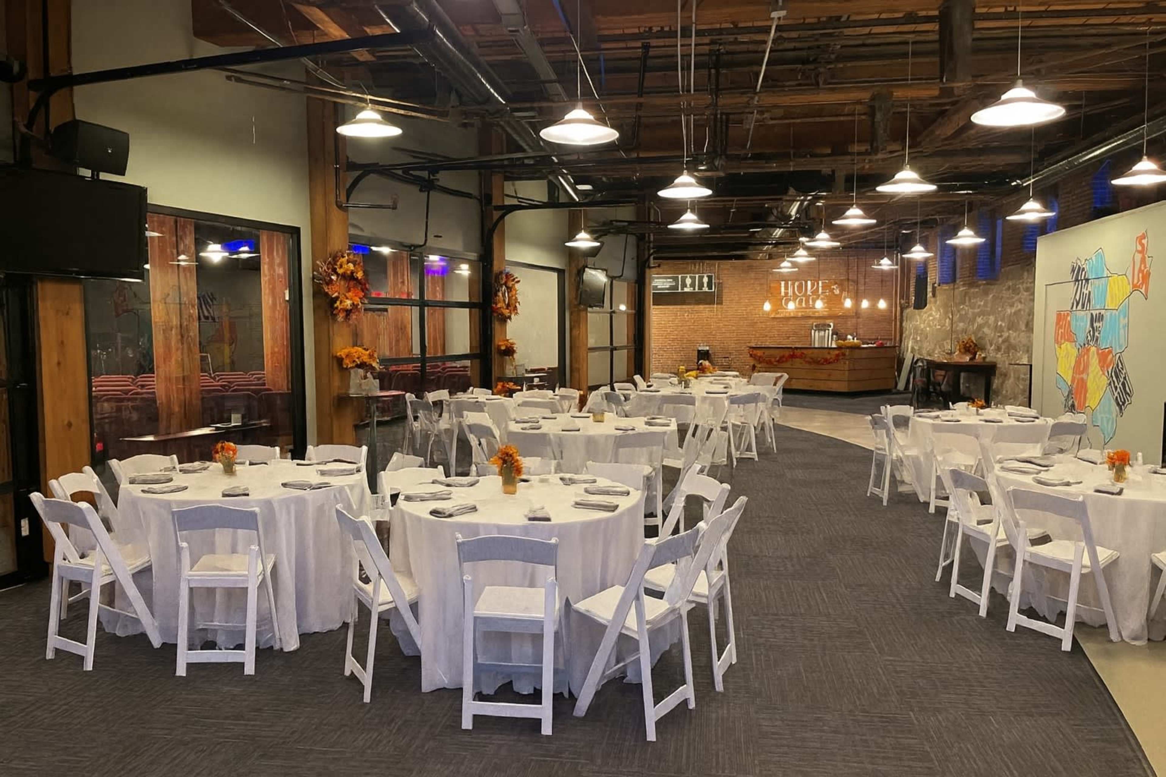 The image shows a dining area set up for an event, featuring multiple round tables covered with white tablecloths and surrounded by white chairs, with autumn decorations and a bar in the background.