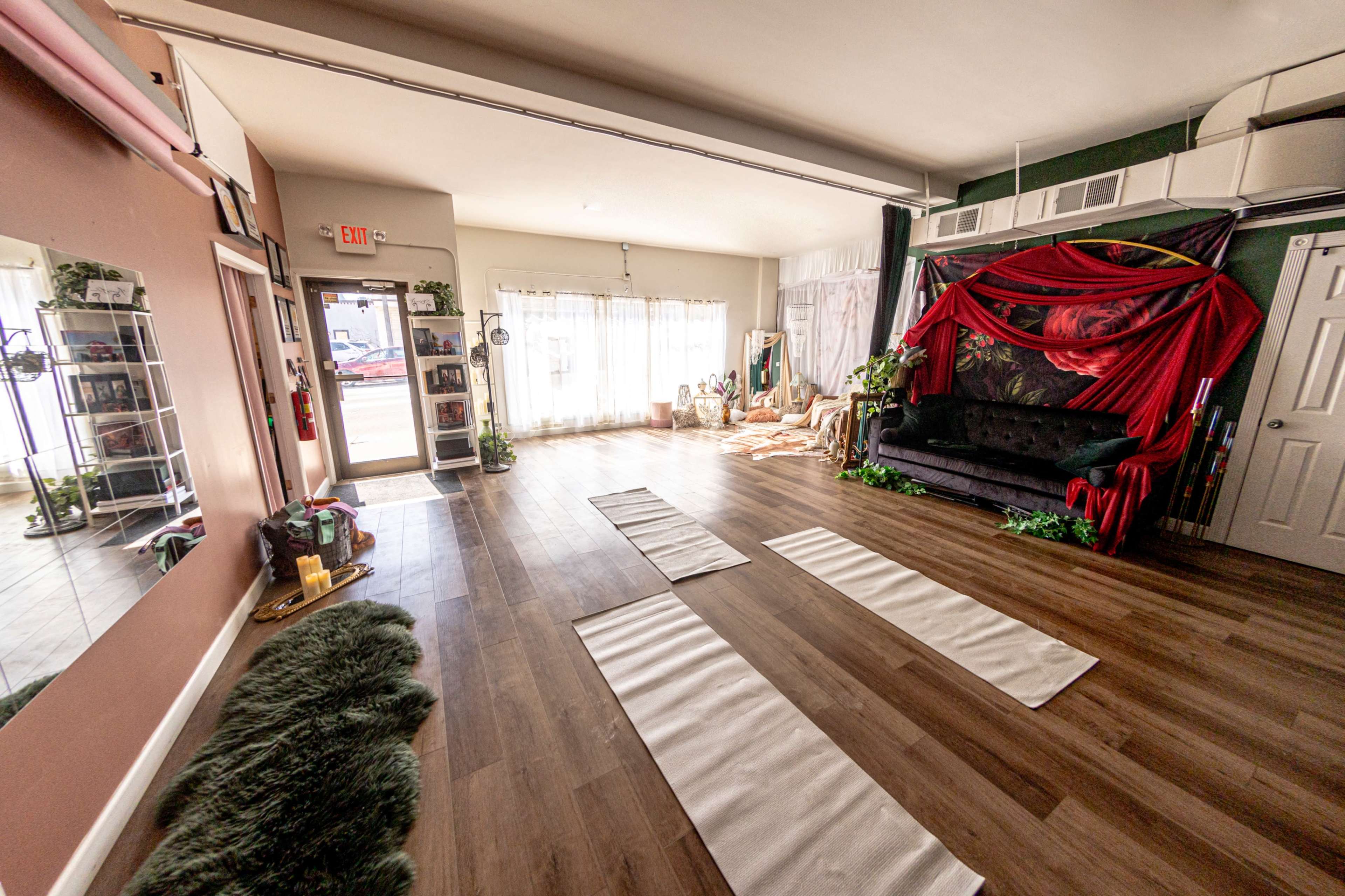 The image shows a spacious yoga studio with wooden flooring, two yoga mats laid out, and a decorative area featuring a black couch and draped fabric.
