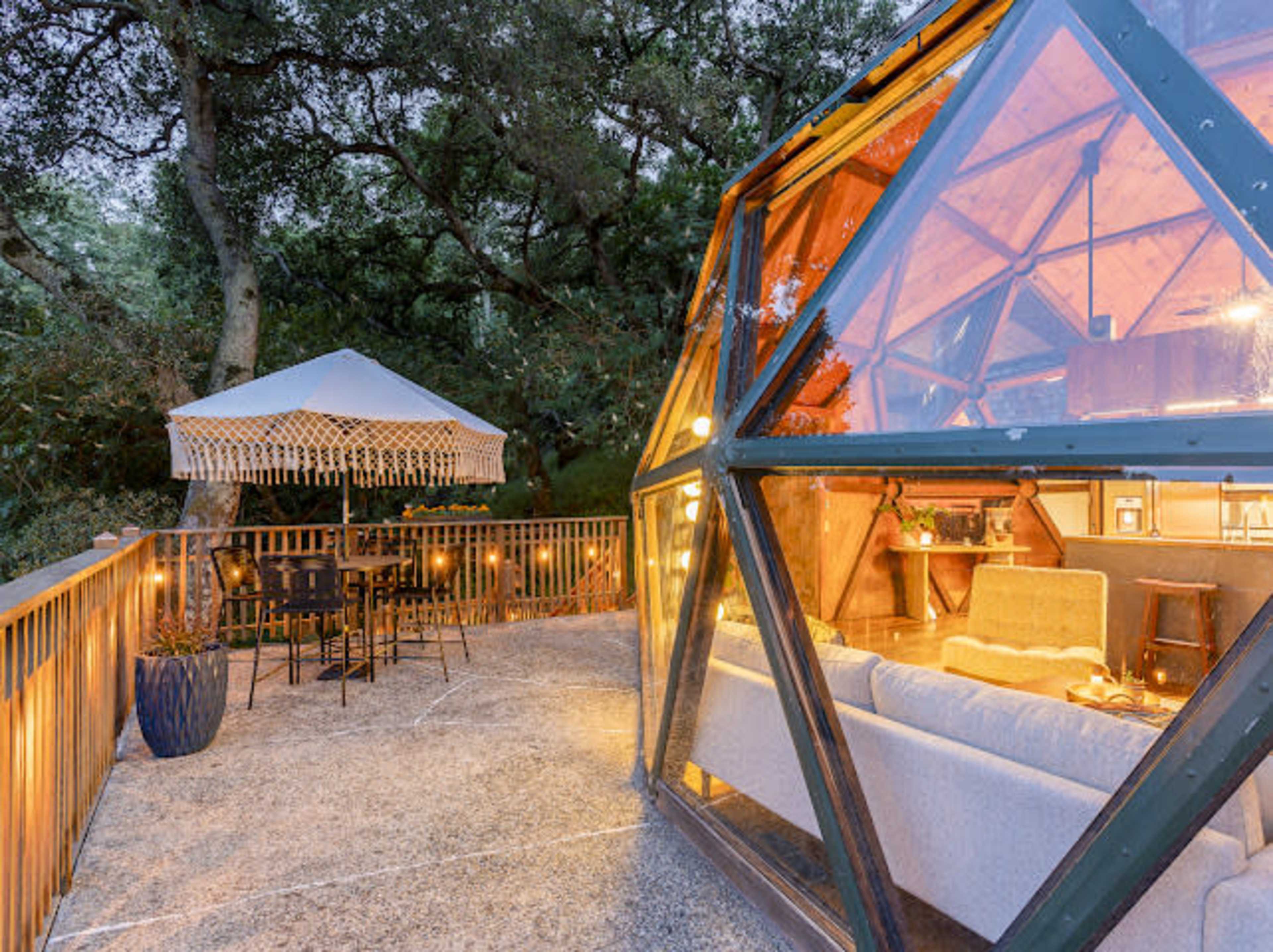 The image shows a modern, geometric glass structure with a cozy interior, surrounded by greenery and featuring an outdoor seating area with string lights and a small gazebo.