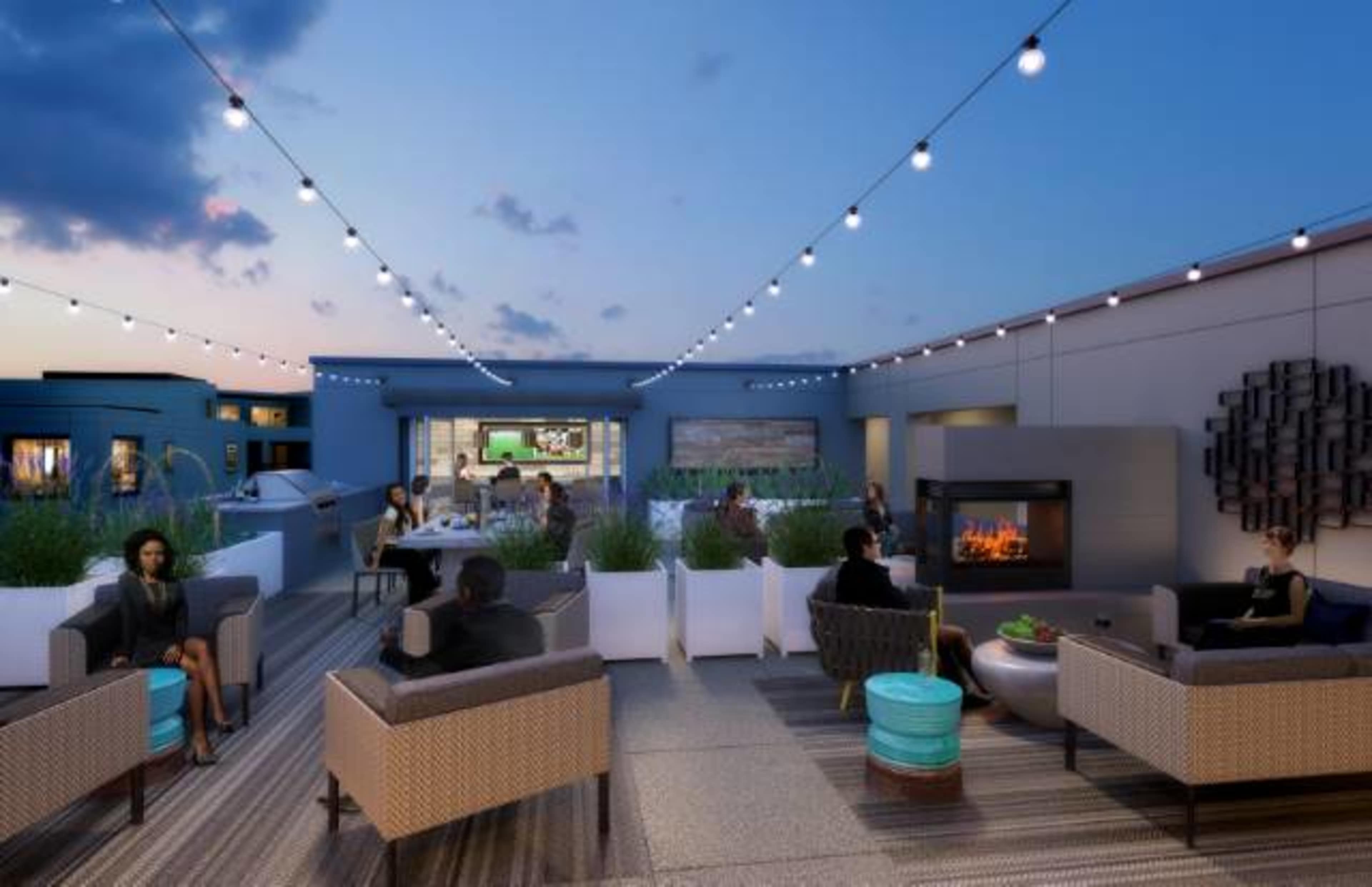 A rooftop lounge area features comfortable seating, string lights, and a modern fireplace, with people socializing in the evening.