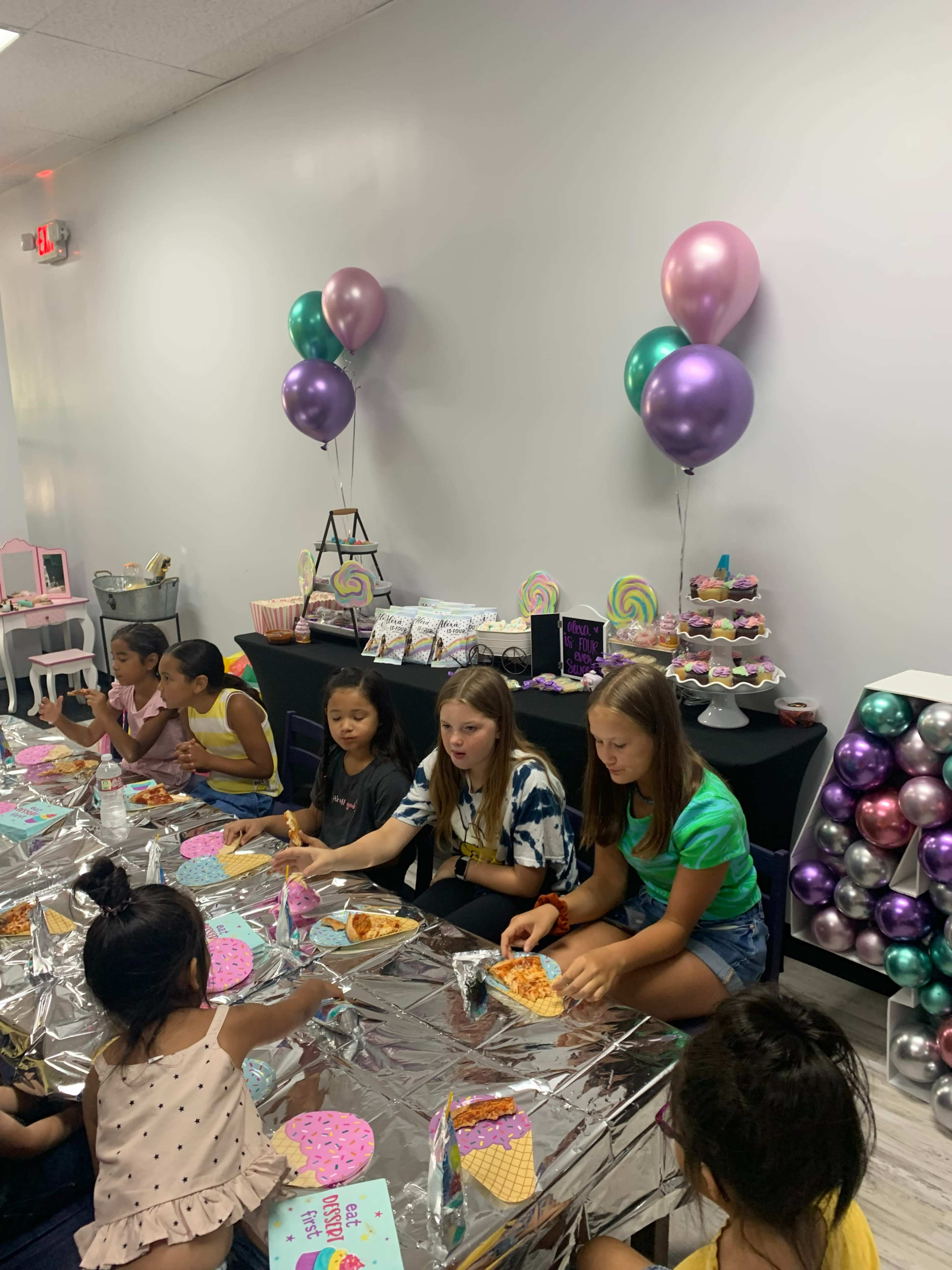 A group of young girls sits at a decorated table, enjoying pizza and snacks at a colorful birthday party.