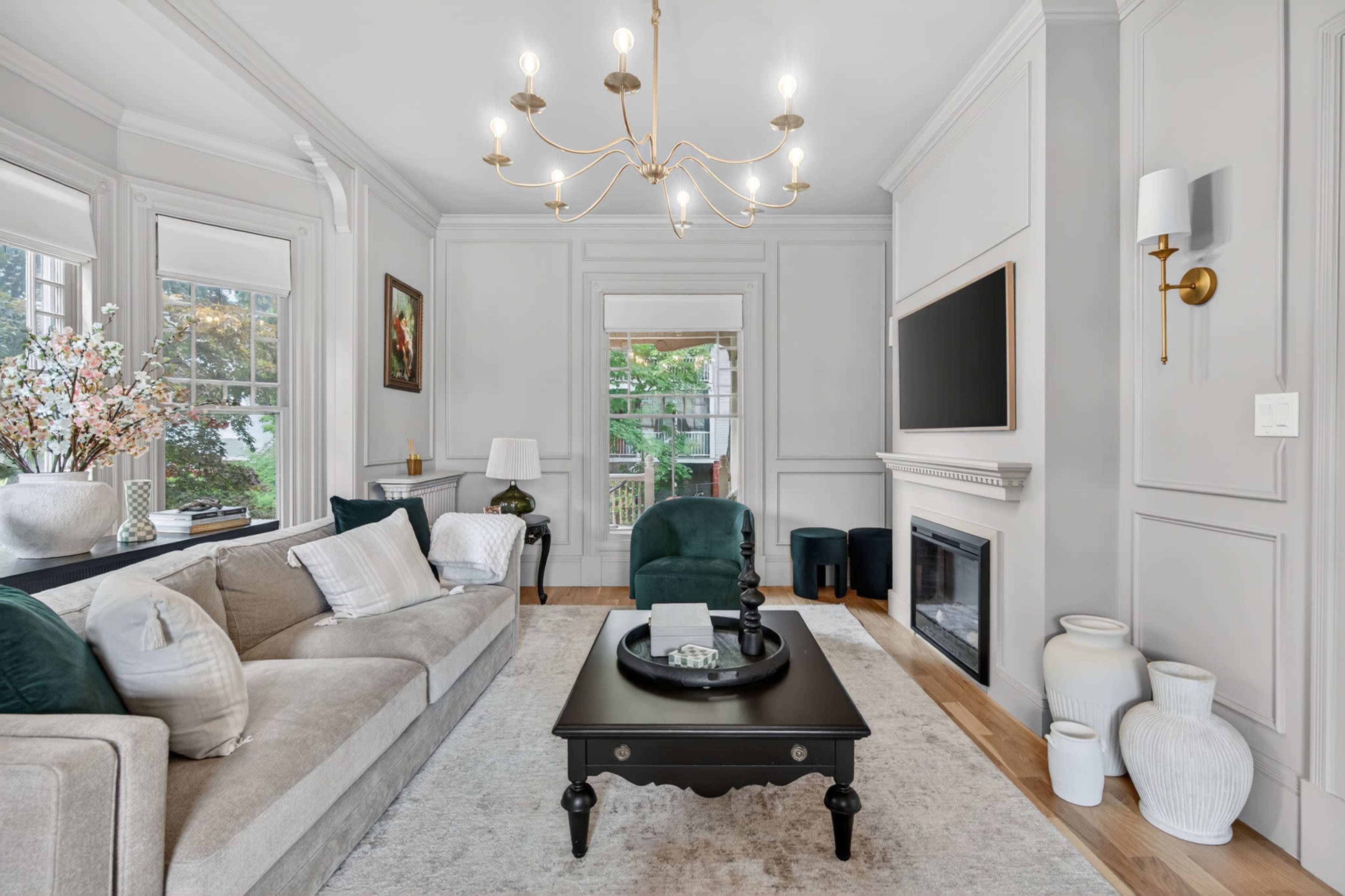 The living room features a light gray sofa, a black coffee table, and a chandelier, with large windows providing views of the outdoors.