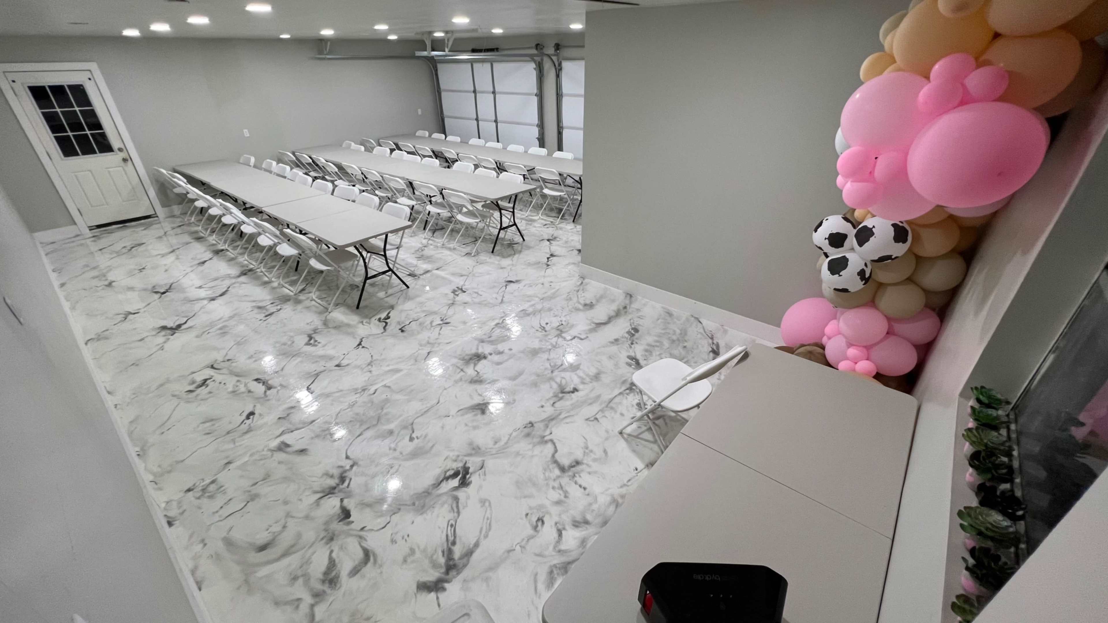 A large room set up for an event, featuring multiple tables arranged in rows, white chairs, and a decorative balloon arrangement in the corner.
