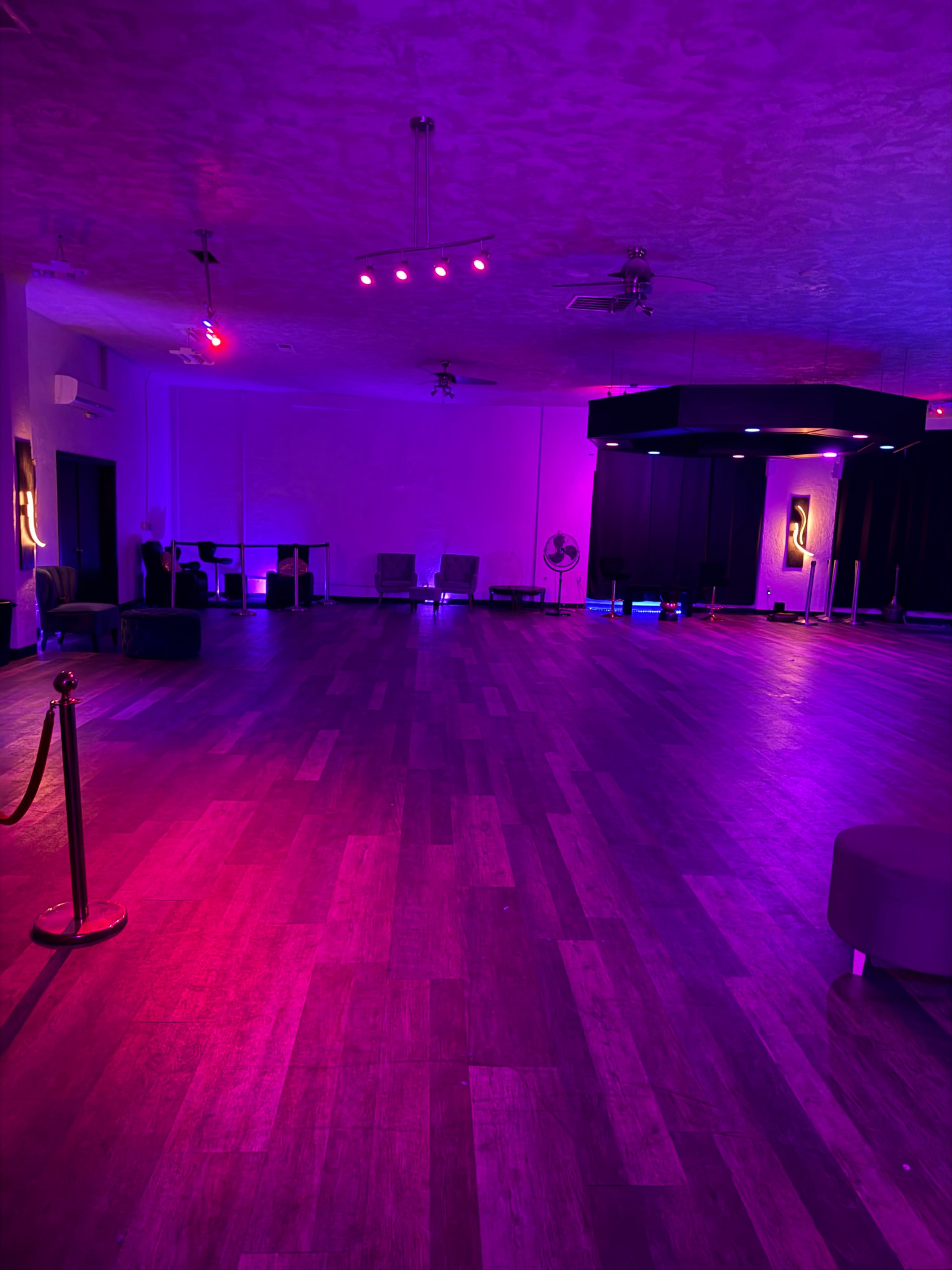 The image shows an empty event space with wooden flooring, illuminated by purple and pink lighting.