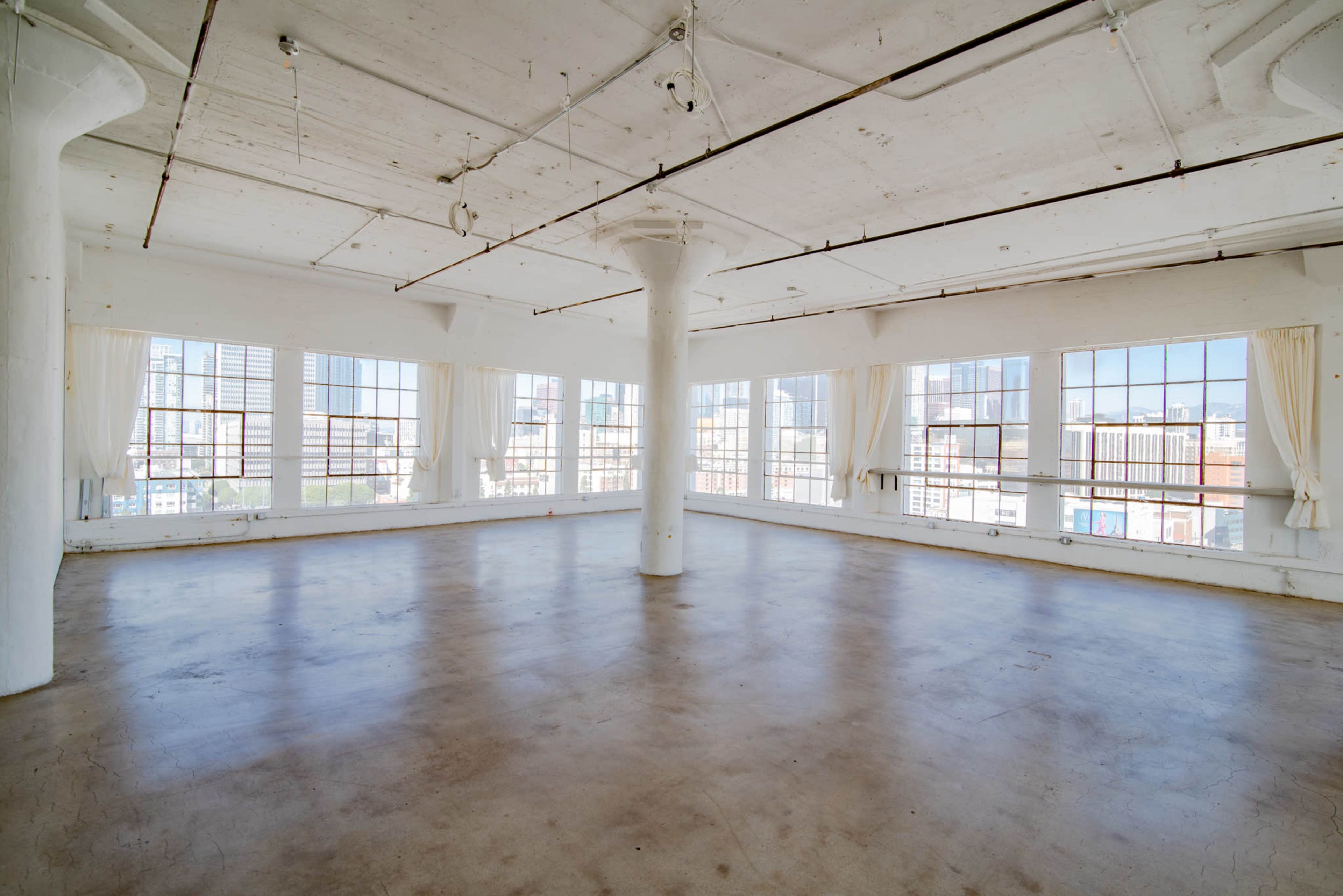 A spacious, empty loft with large windows and concrete pillars, bathed in natural light.