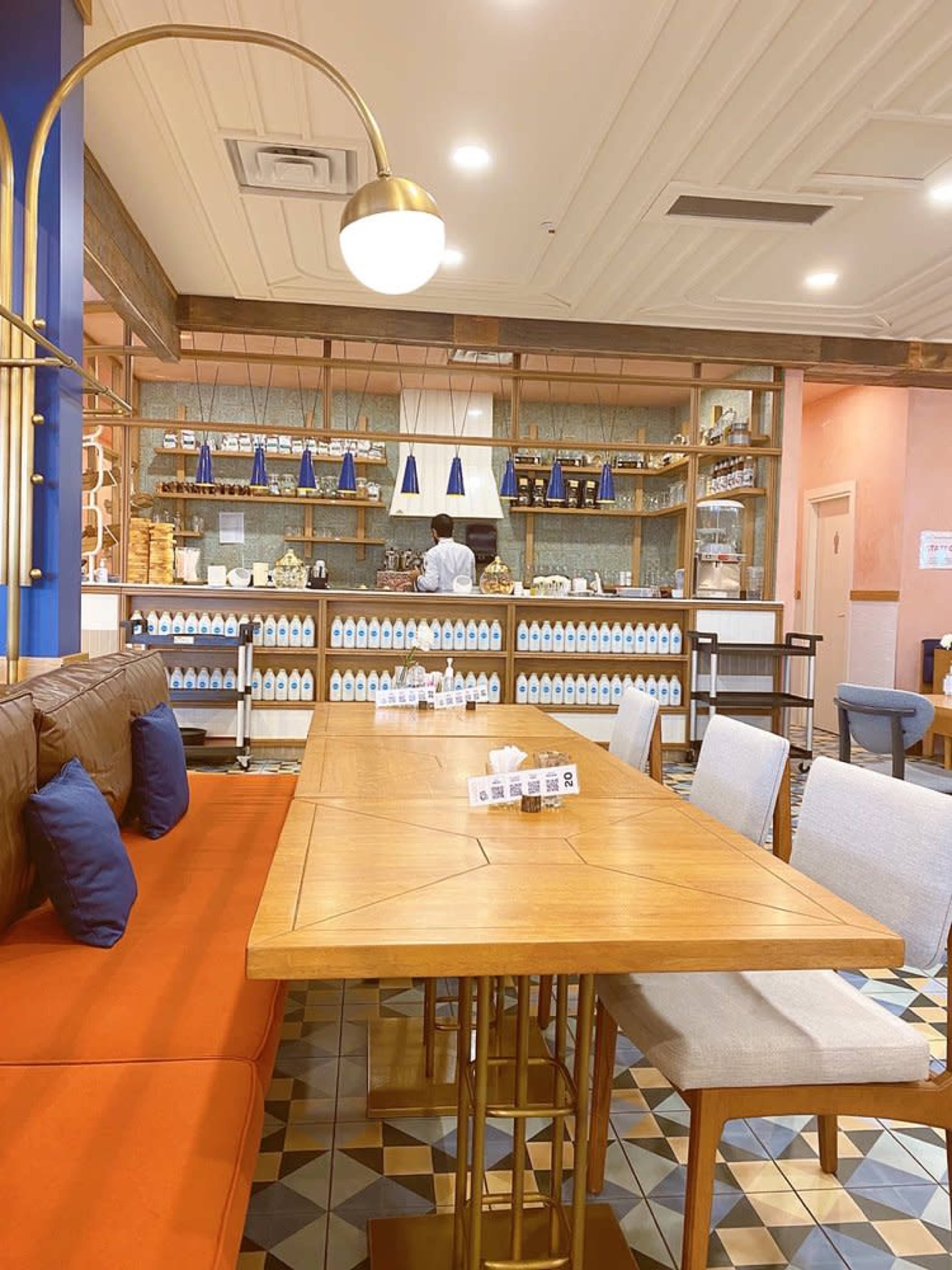 A cozy cafe features a wooden table with orange and gray seating, shelves stocked with bottles, and a staff member behind the counter.