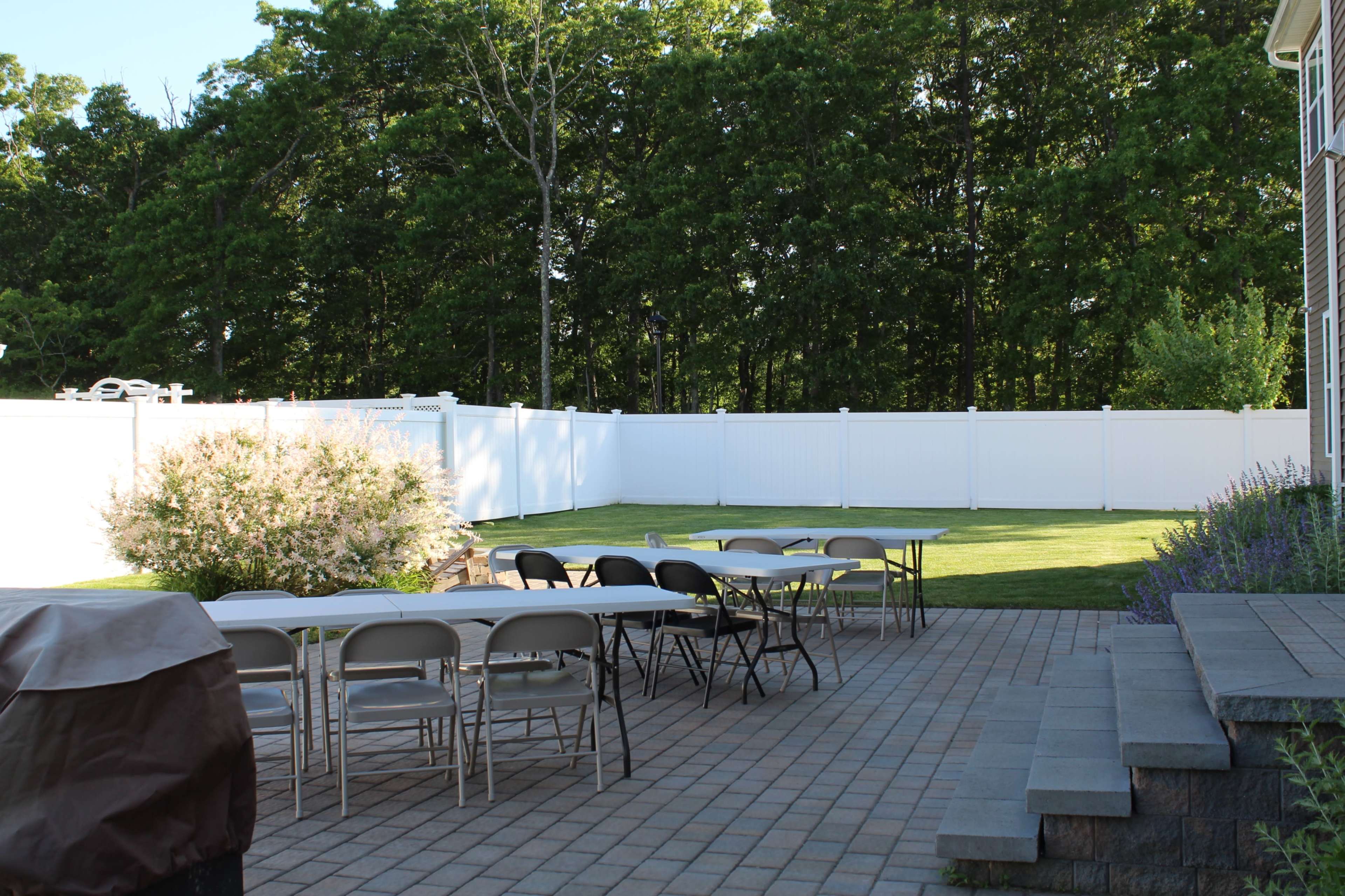A neatly arranged outdoor space features several tables and chairs on a patio near a grass lawn, surrounded by a white fence and trees.