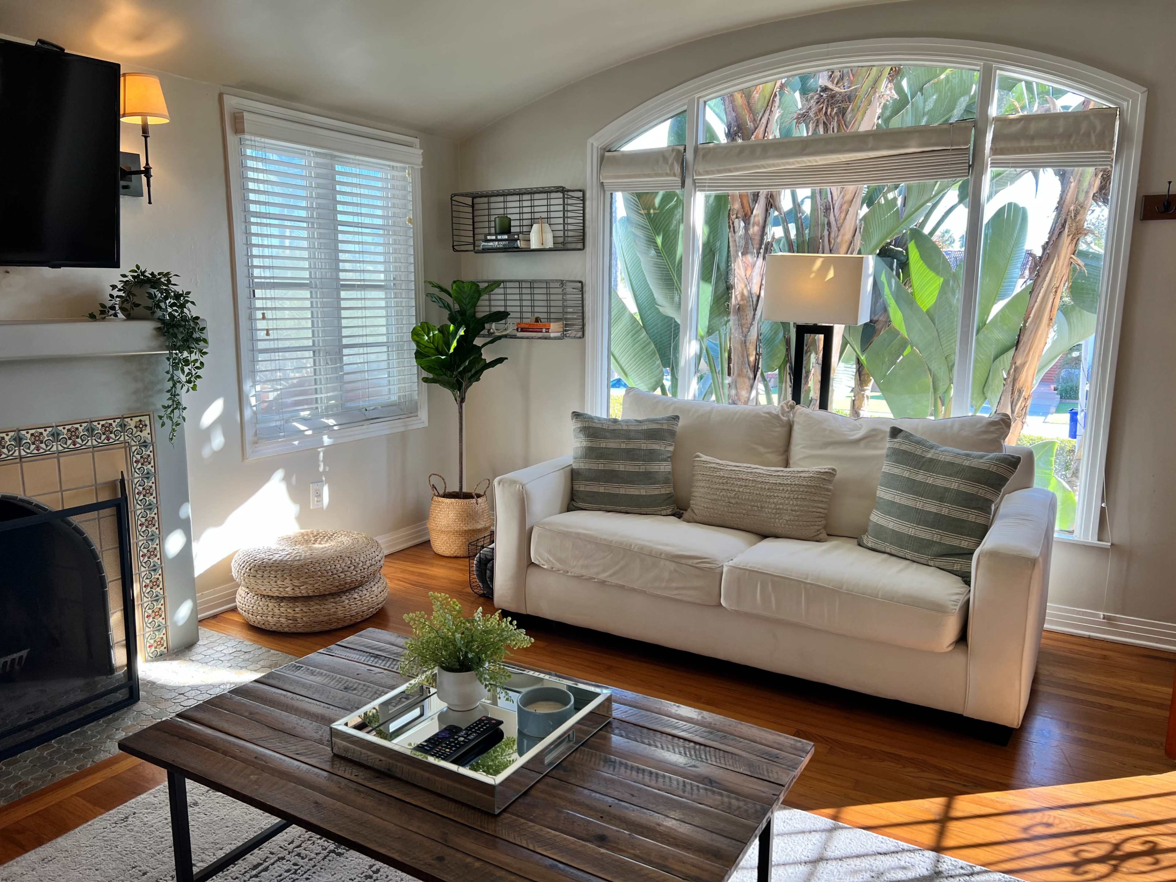 The image shows a cozy living room featuring a white sofa, a wooden coffee table with a small plant, and large windows that provide natural light and a view of greenery outside.