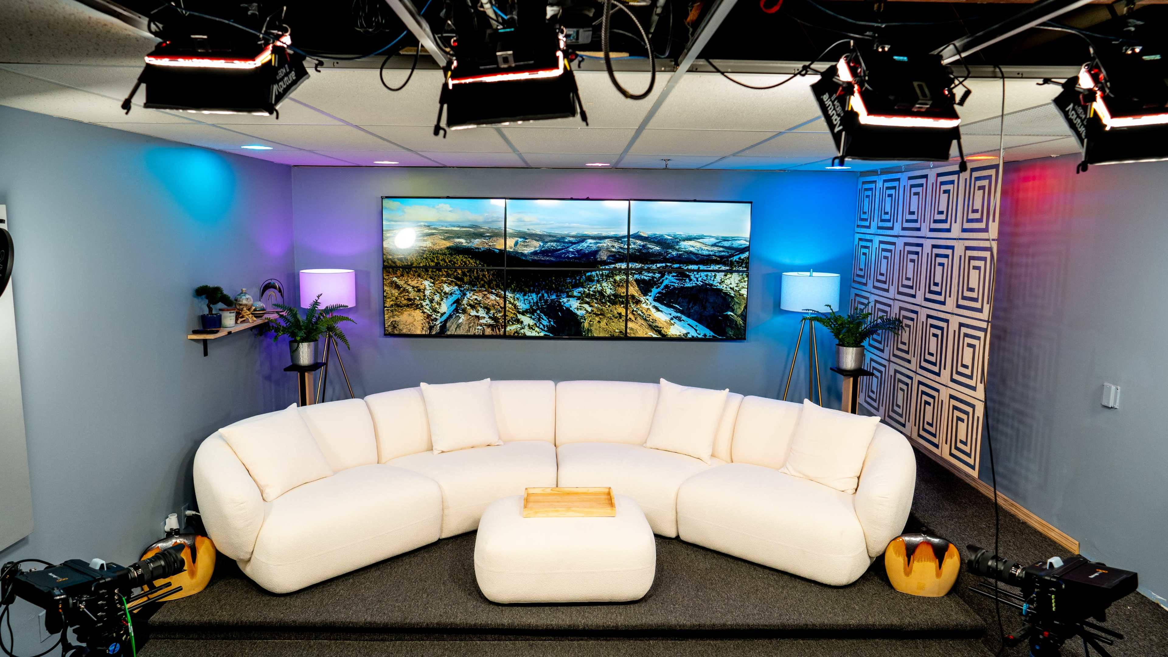 A modern, circular white sofa in a studio setting, with a large screen displaying a snowy mountain landscape in the background and studio lights overhead.