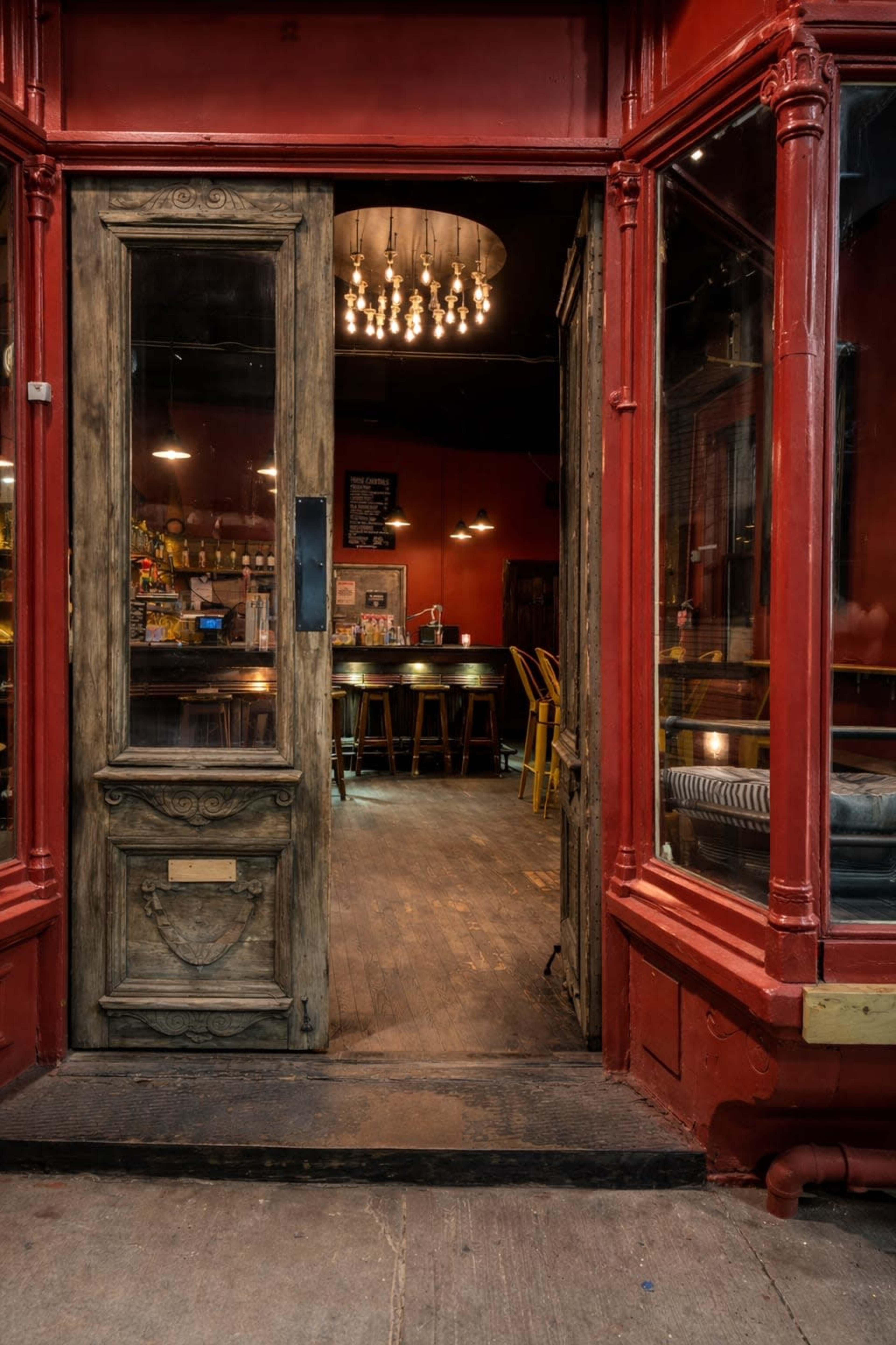 The image shows the entrance of a restaurant with a wooden door and a red exterior, leading into a dimly lit interior featuring tables and hanging lights.