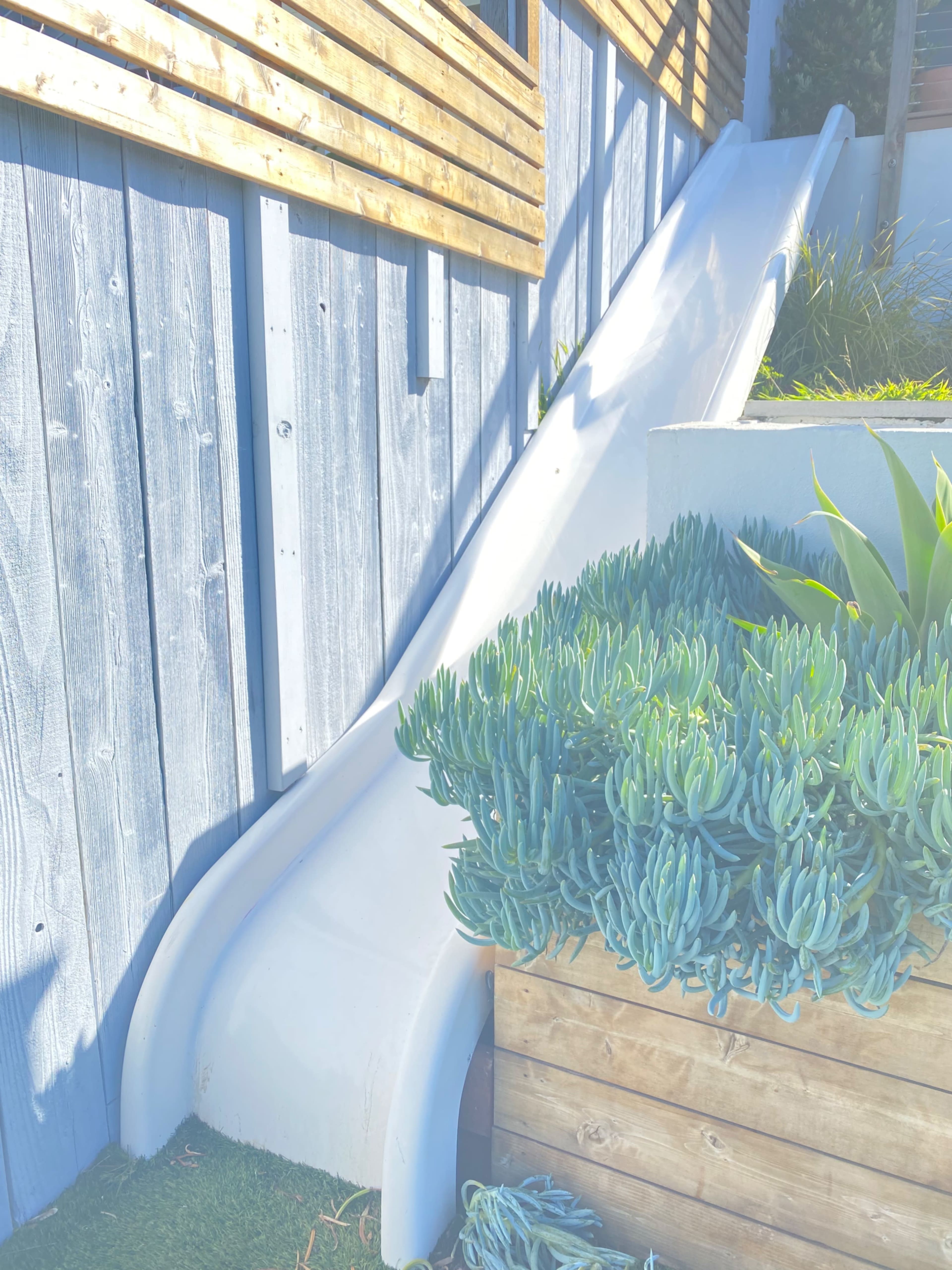 The image shows a white slide positioned next to a wooden wall and surrounded by green plants in a landscaped area.