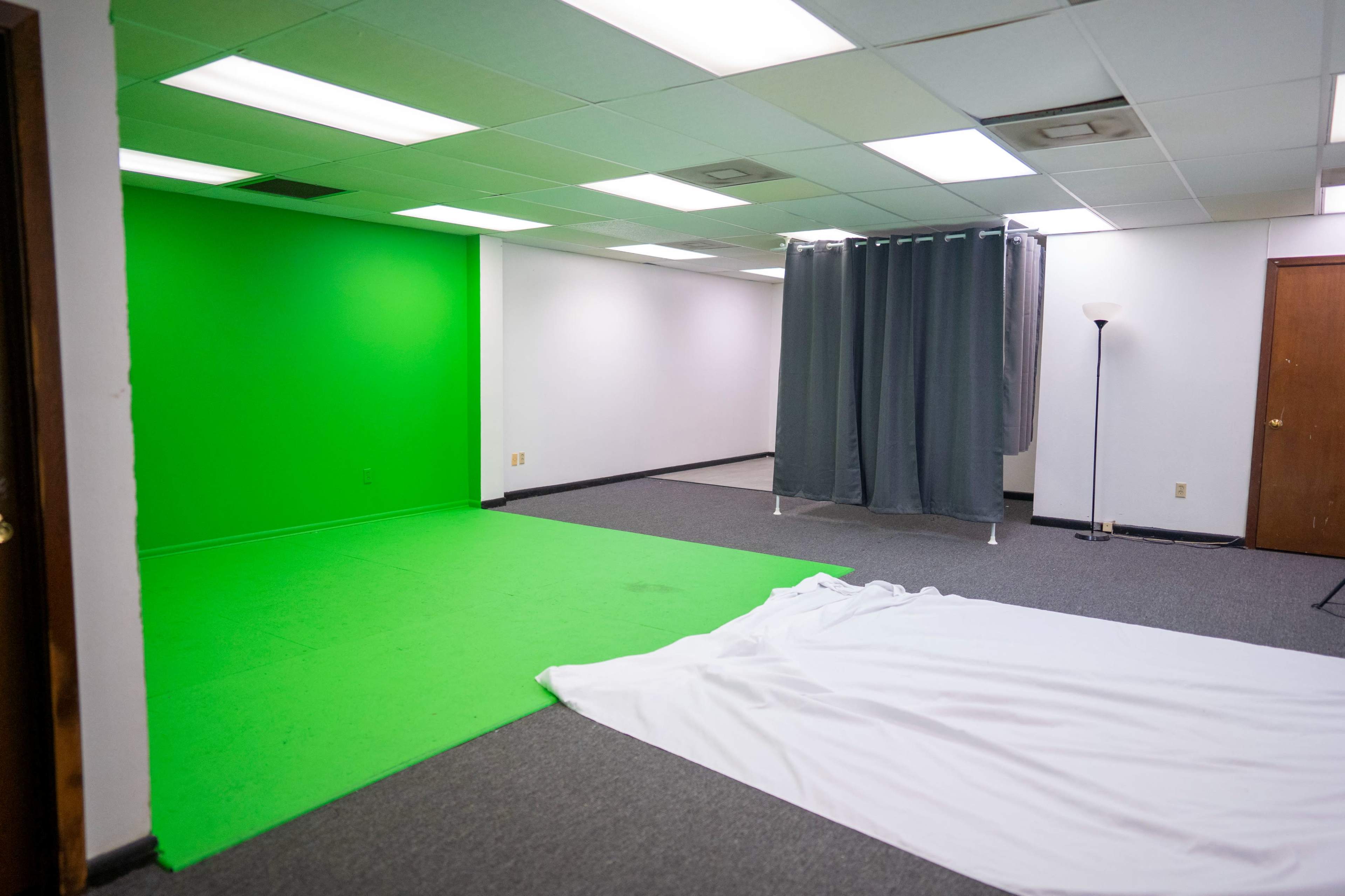 The image shows a room with a bright green screen on one side and a white fabric laid out on the carpeted floor, while gray curtains are partially closed in the corner.