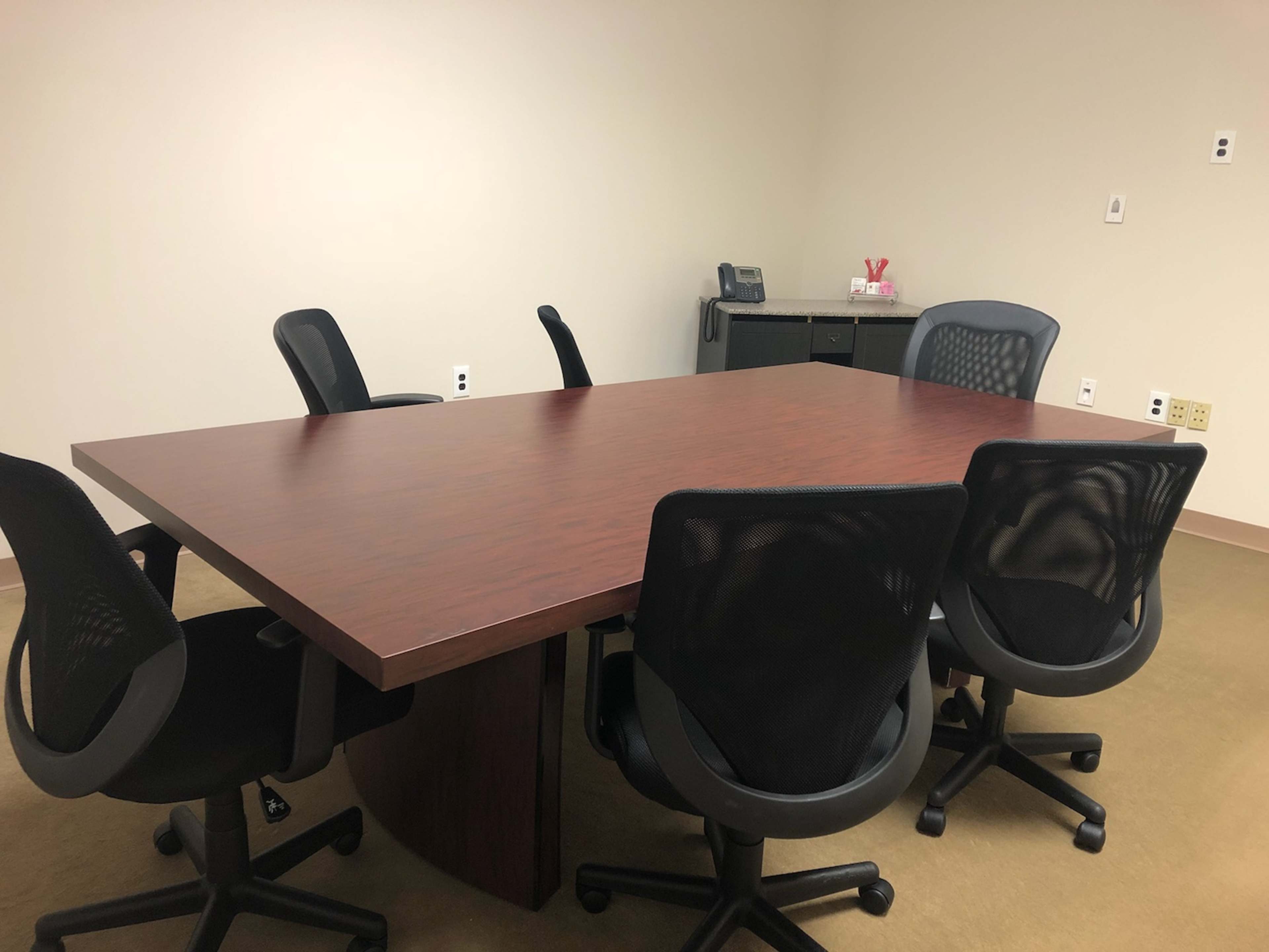 A simple conference room features a large wooden table surrounded by five office chairs, with a telephone and a cabinet visible in the background.